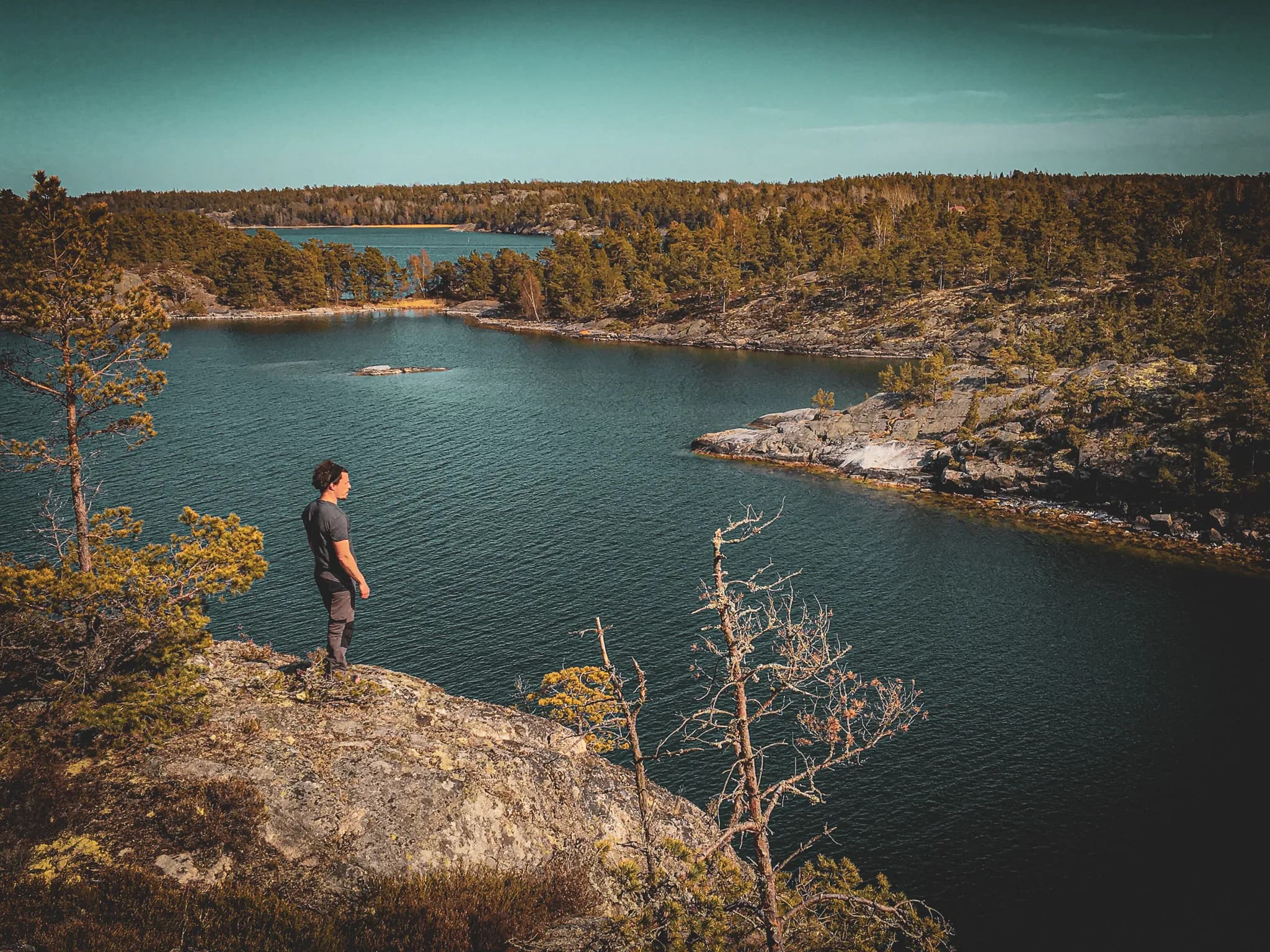 A man stands on the edge of a rocky cliff overlooking a calm body of water, surrounded by wooded landscapes. In the distance, several islands can be seen in the lake, while the vegetation, made up of