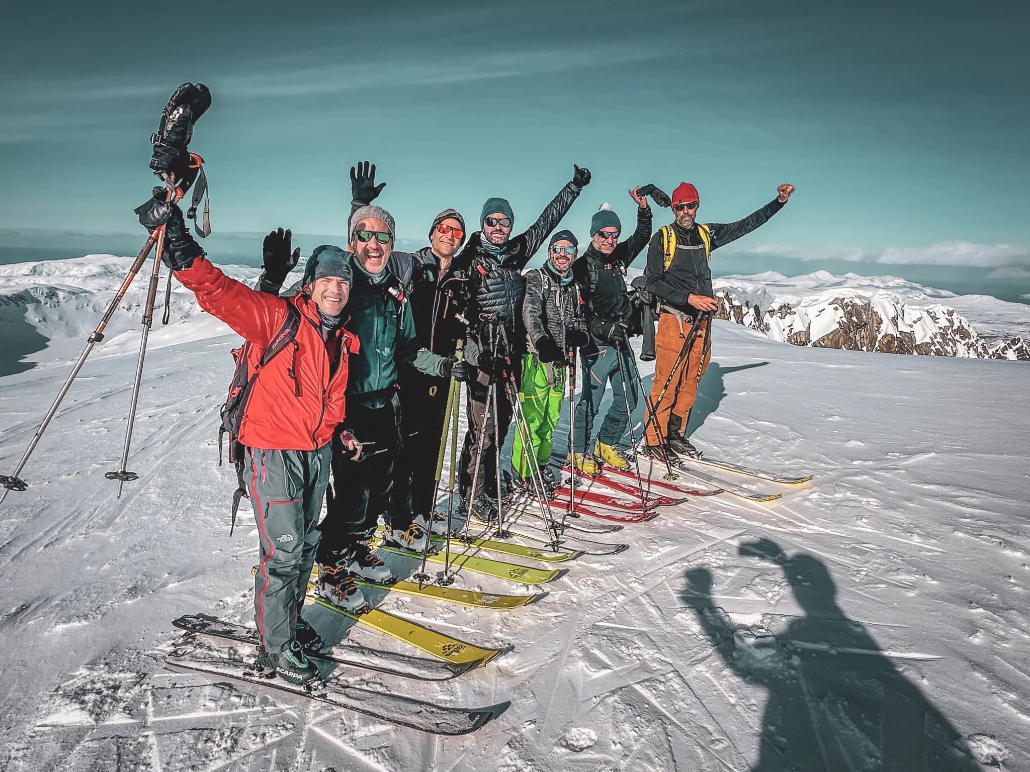 Un groupe de skieurs posant sur un sommet enneigé, tout sourire et visiblement ravis. Les membres du groupe portent des vêtements colorés adaptés au ski, et certains lèvent les bras en signe de célébration. La neige gl