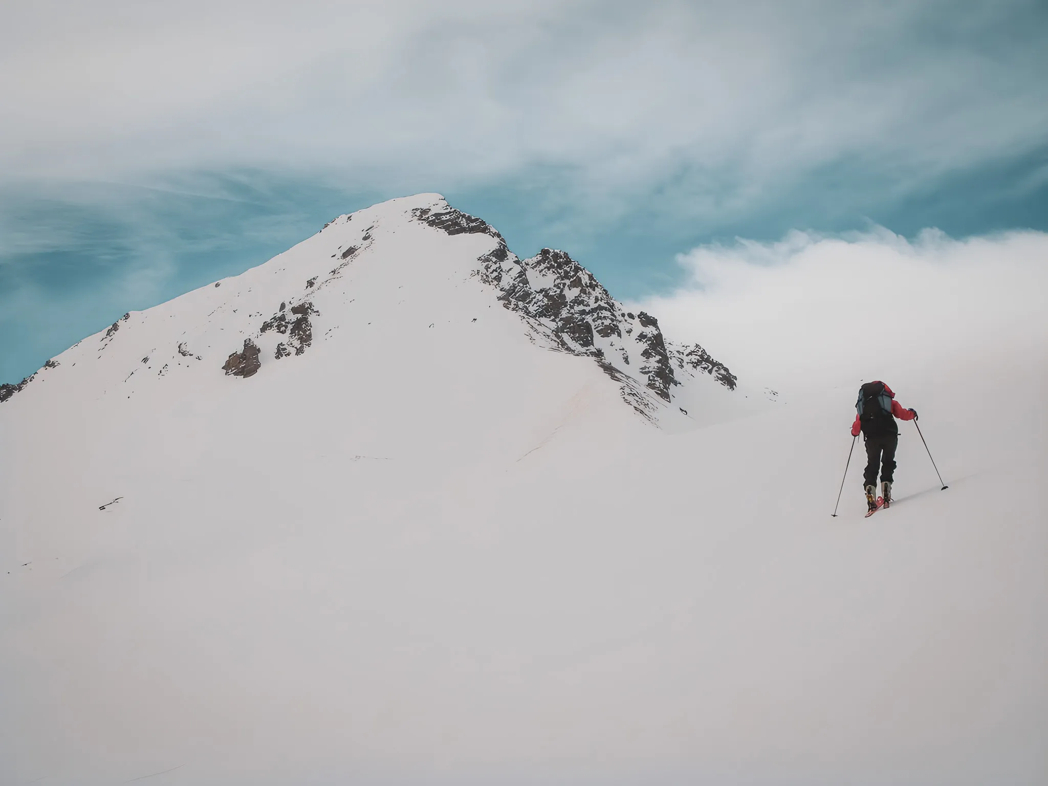 A skier ascends a snow-covered slope, facing majestic peaks under a blue sky.