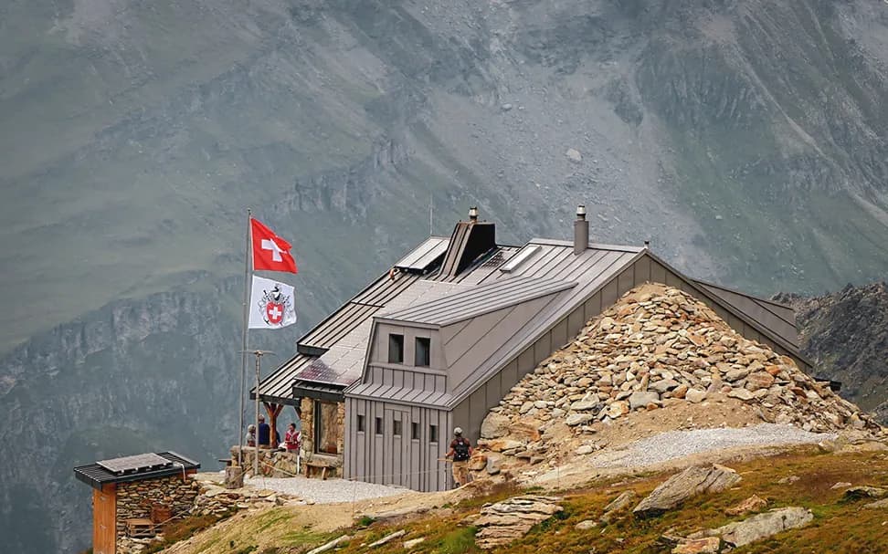 A modern building with a sloping roof is situated high up, surrounded by mountains. Two flags are visible: one is the Swiss flag, and the other bears a coat of arms.