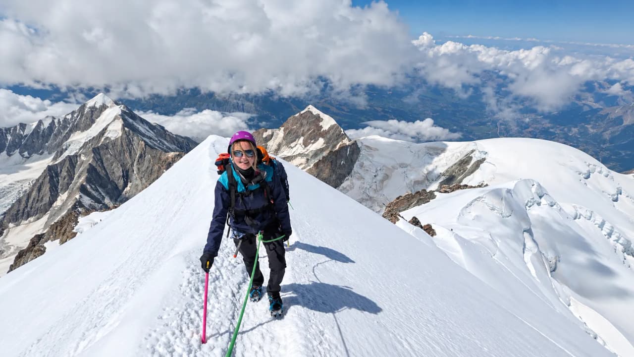 Alpiniste souriante sur un sommet enneigé, entourée de chaînes de montagnes majestueuses.