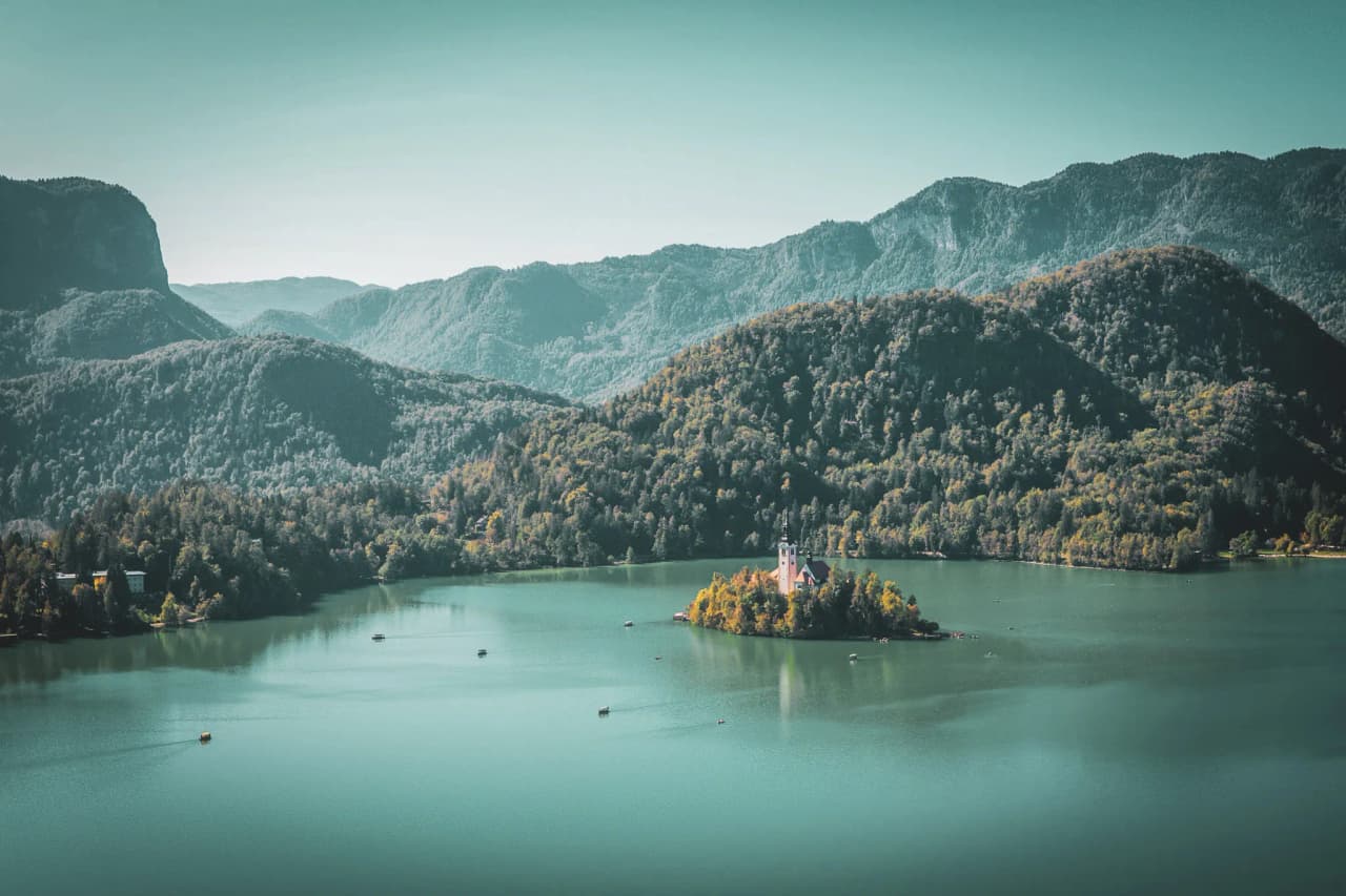A tranquil lake surrounded by green mountains, with a small island in the centre where a charming church stands. The landscape is bathed in soft light, and small boats sail peacefully on the water.