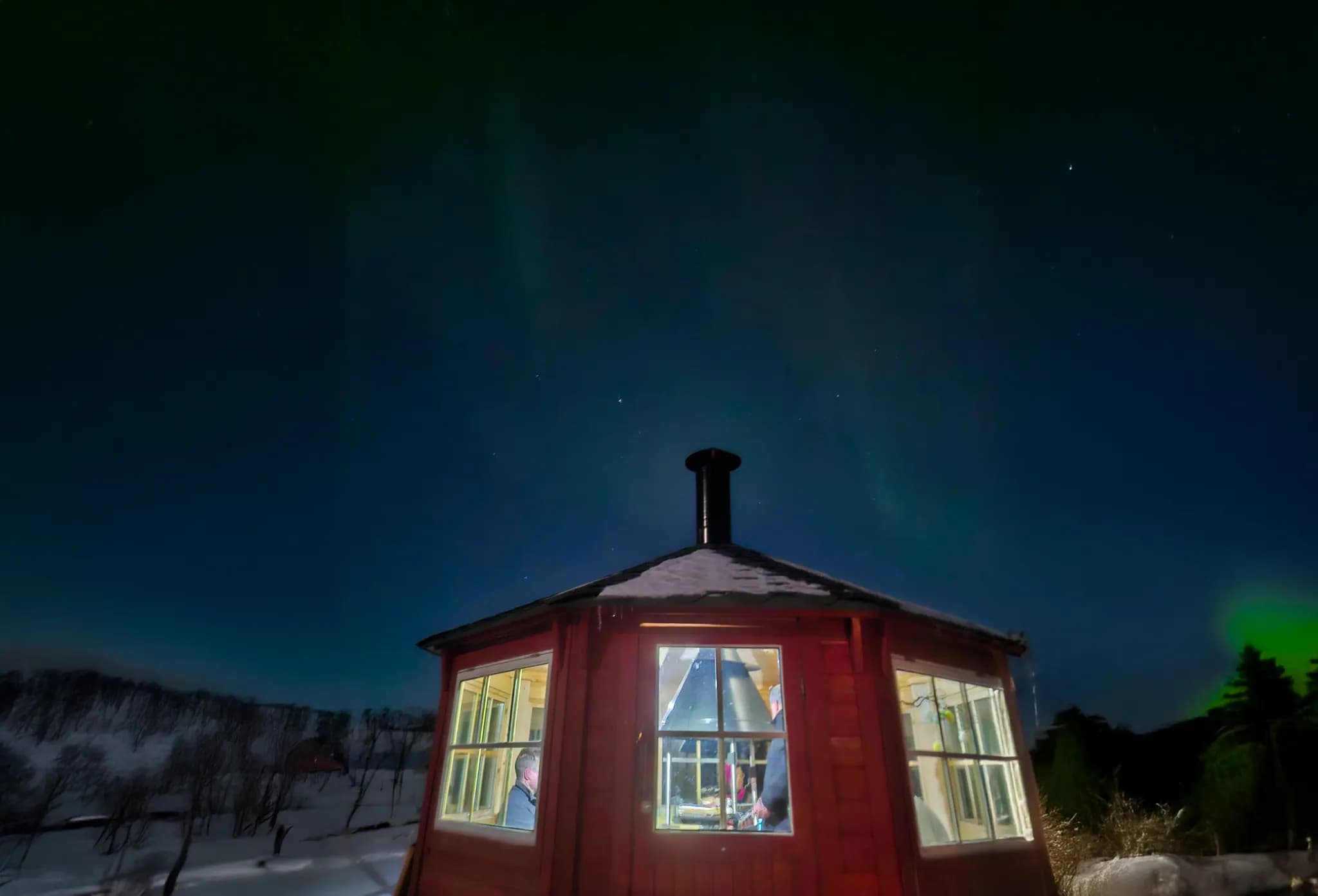 A red wooden chalet with large windows, bathed in a nocturnal atmosphere, under a starry sky. Snow covers the ground all around, and a chimney emerges from the roof. Inside, two people are visible,