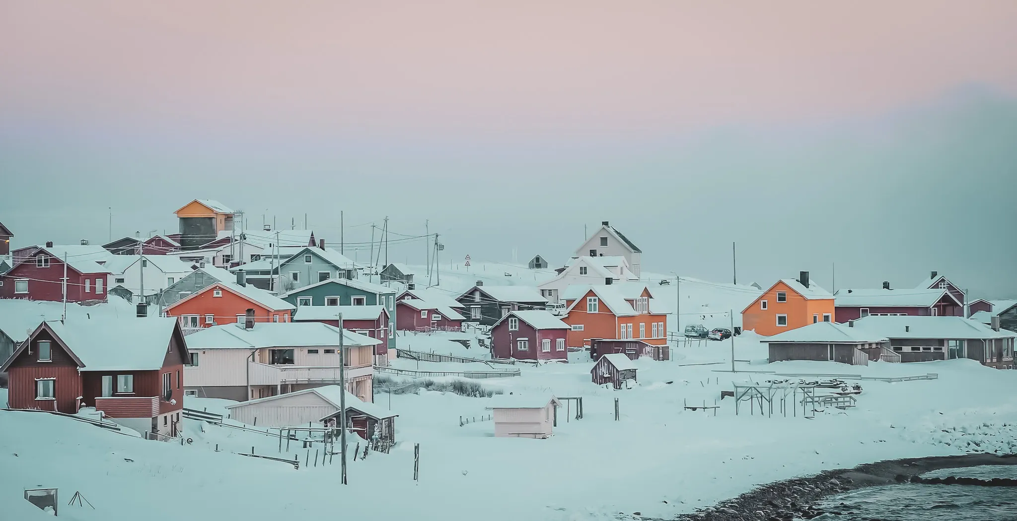 A colourful snowy village, where houses in bright shades of orange, red and green contrast with the winter landscape. The roofs of the buildings are covered in snow, and some structures have
