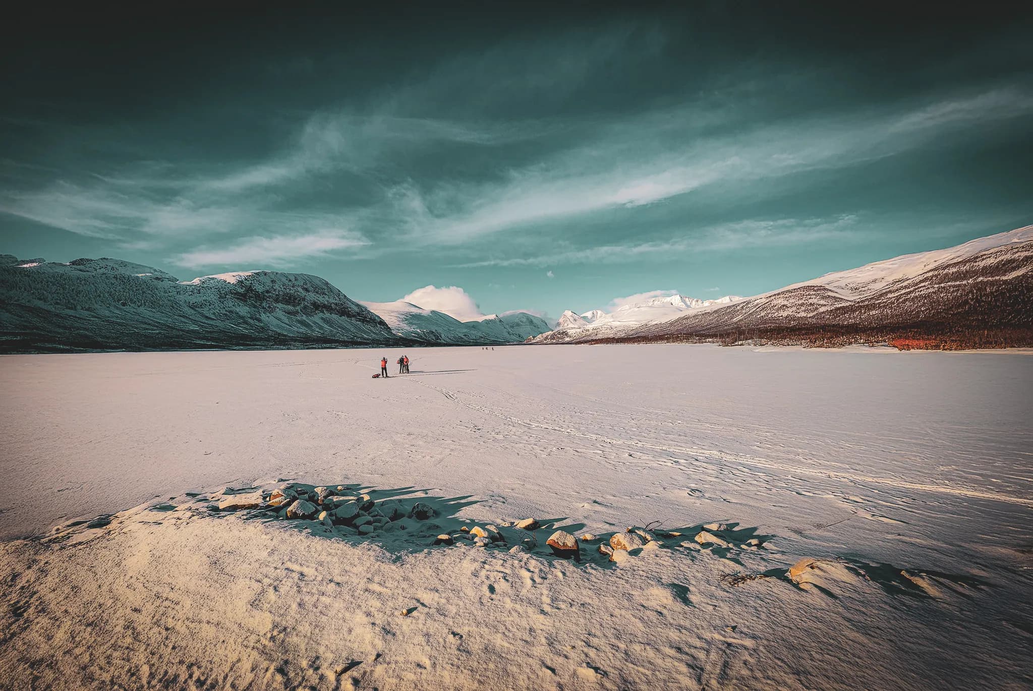 A vast, snow-covered Arctic landscape, with mountains in the distance under a slightly cloudy sky. In the foreground, a group of people on snowshoes move across the ice, dragging equipment with a pulka.