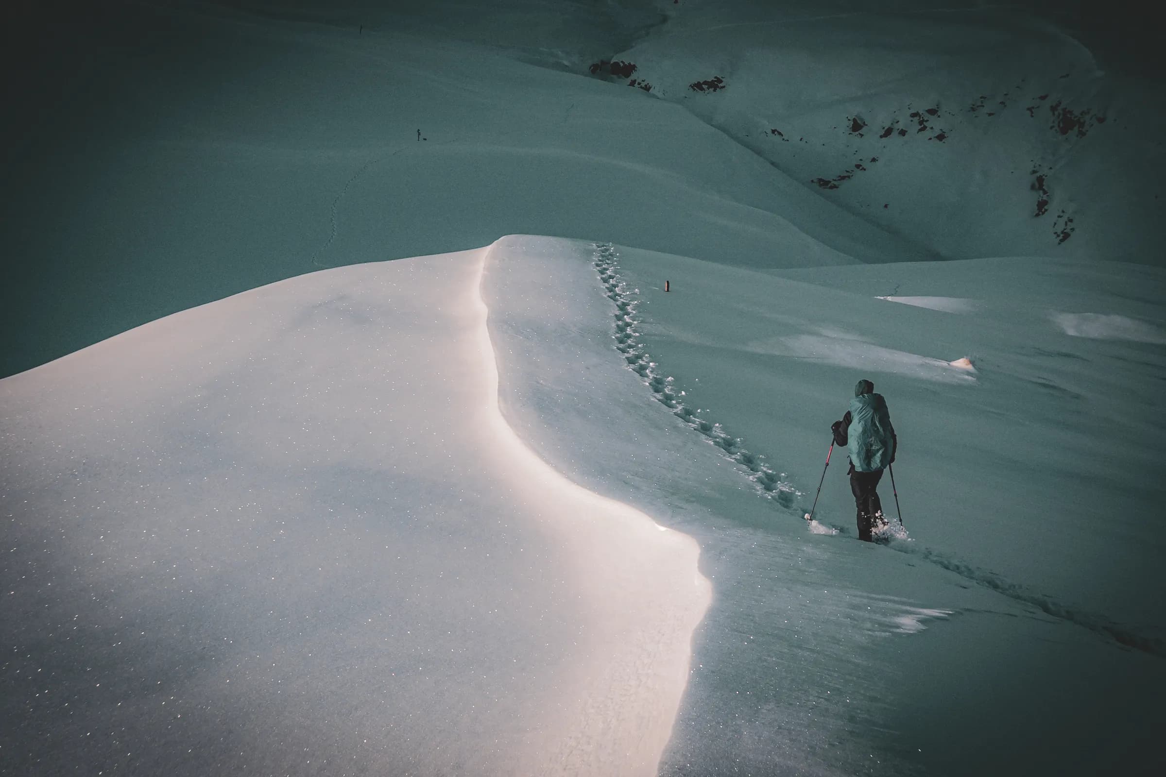 A hiker walks across a vast snow-covered landscape, with fresh footprints left in the glistening snow. The silhouette of the walker, equipped with hiking poles and a rucksack, comes into view.