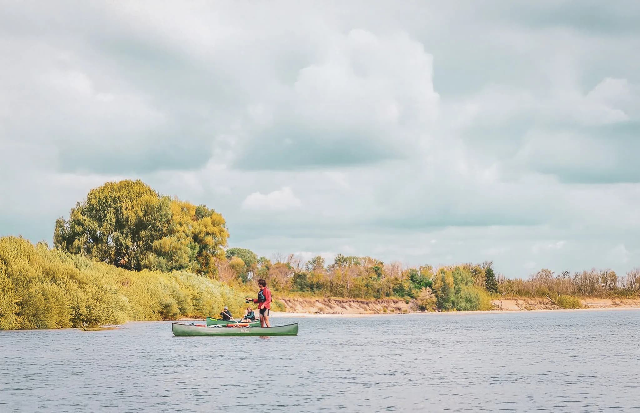 A canoe close to a green bank, with several people on board. The sky is cloudy, creating a peaceful atmosphere. The vegetation on the riverbank is lush, with trees with blazing leaves.