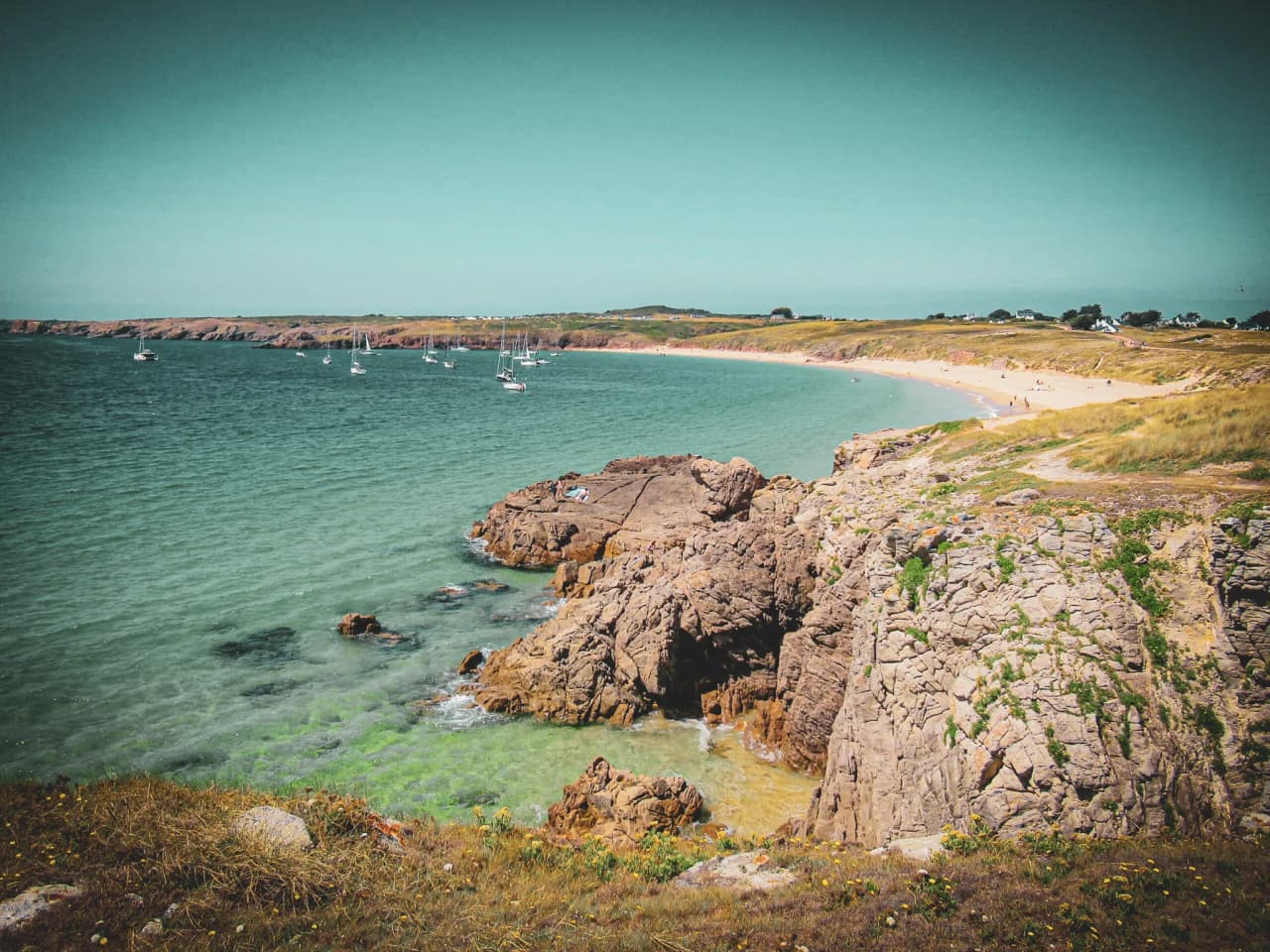 Goudkleurig zandstrand omzoomd door rotsen, zeilboten in de verte, blauwe luchten boven de wilde Bretonse kust.