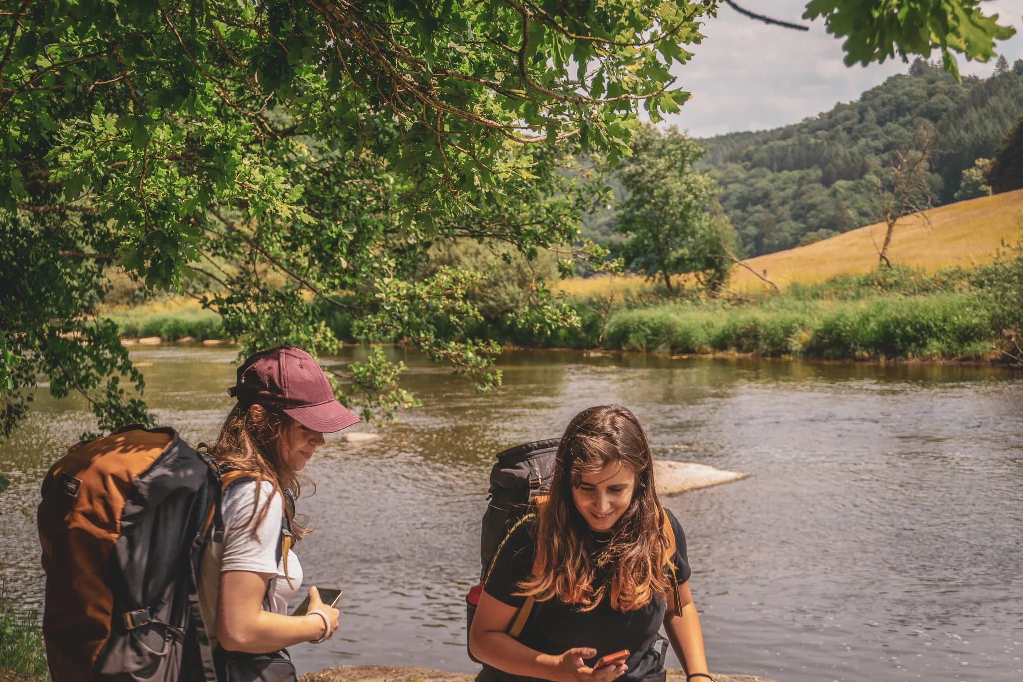Twee vrouwen met grote rugzakken staan bij een rivier, omringd door weelderig groen. Een van hen, met een pet op, kijkt naar haar telefoon, terwijl de ander glimlacht en geïnteresseerd lijkt in