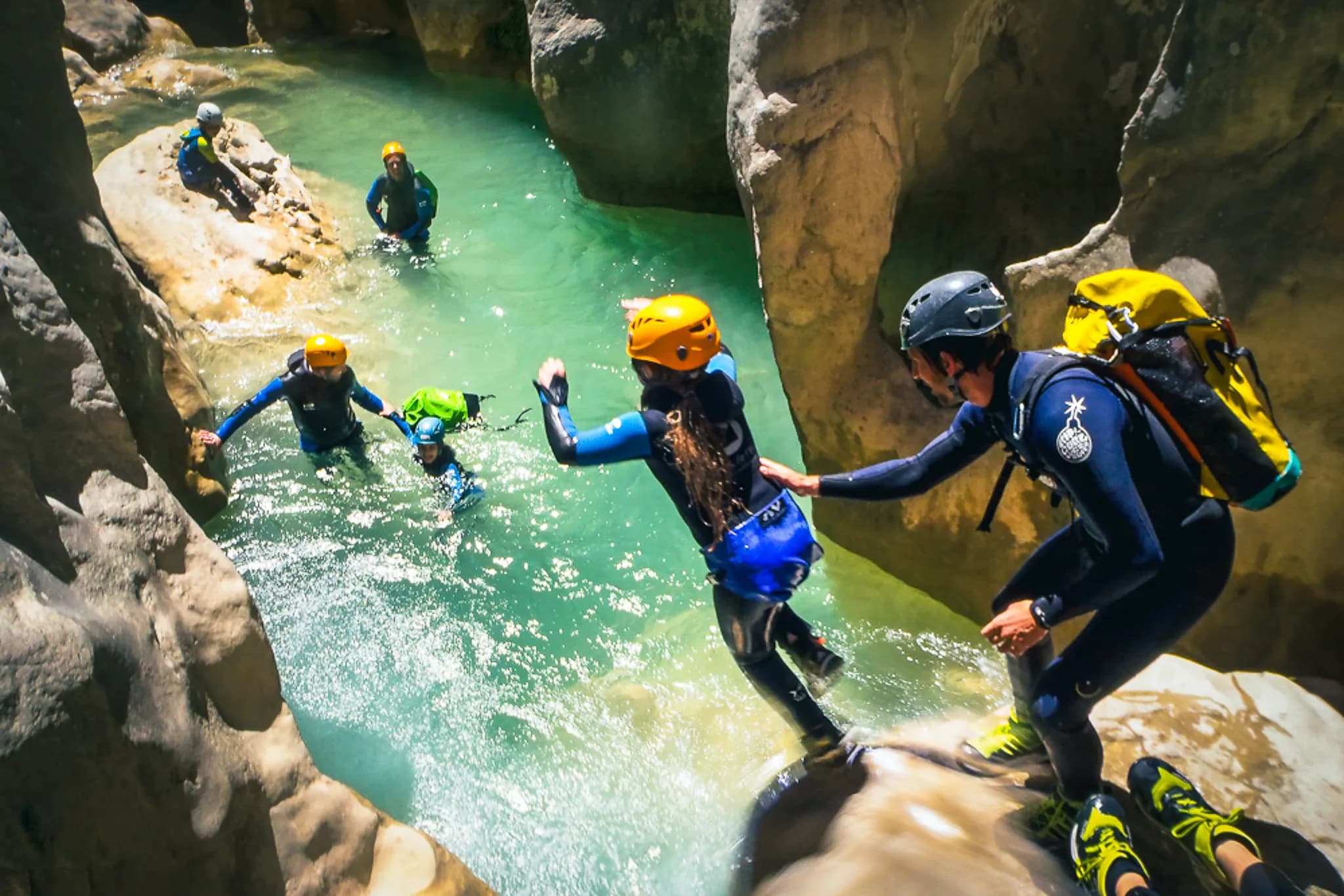 A group of people embark on a water-climbing activity in a canyon. The water is clear and emerald green, surrounded by rock faces. A man helps a woman, who is about to