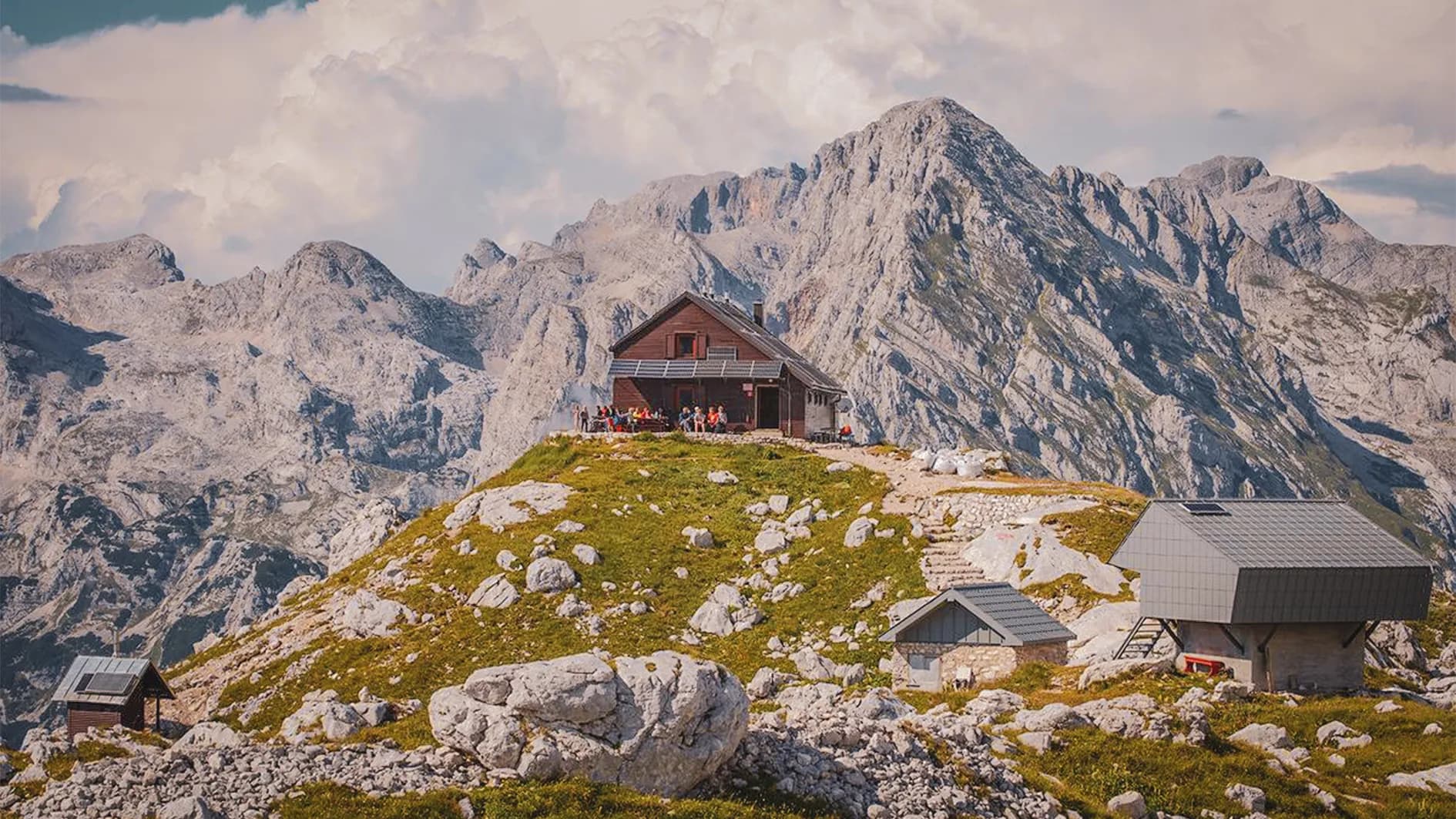 A wooden chalet is set on a green hillside, surrounded by rocks and majestic mountains in the background. The chalet is alive with a group of people gathered on the terrace, enjoying the fresh air.