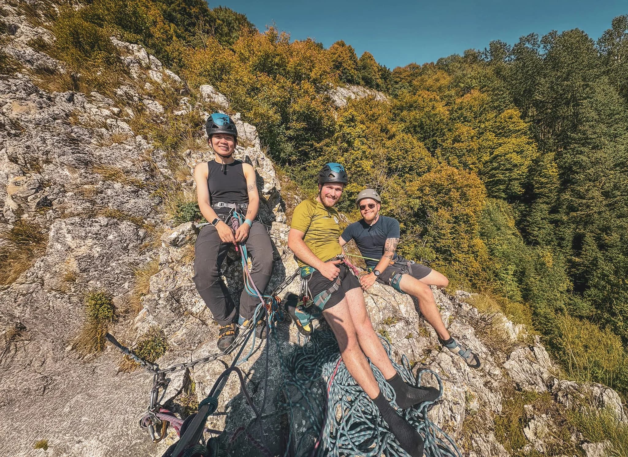 Three smiling climbers rest on a sunny cliff, surrounded by lush nature.