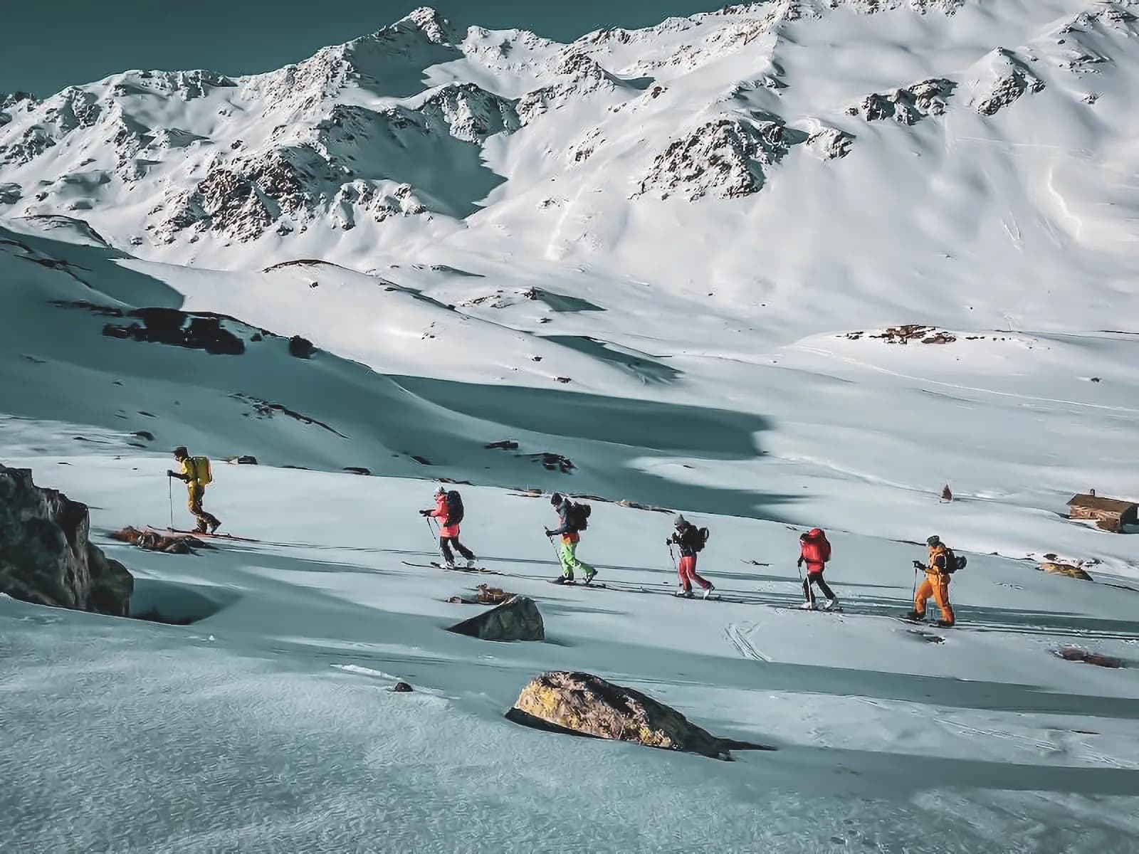 Un groupe de skieurs évolue sur un paysage montagneux enneigé, avec des sommets imposants en arrière-plan. Les skieurs portent des vêtements de couleurs vives, ce qui contraste avec la blancheur de la neige. Le terrain