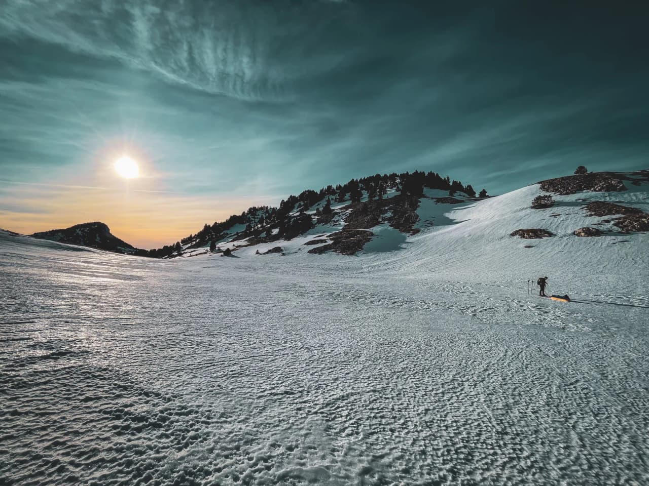 A hiker on snowshoes makes his way across a snow-covered landscape, under a majestic setting sun.