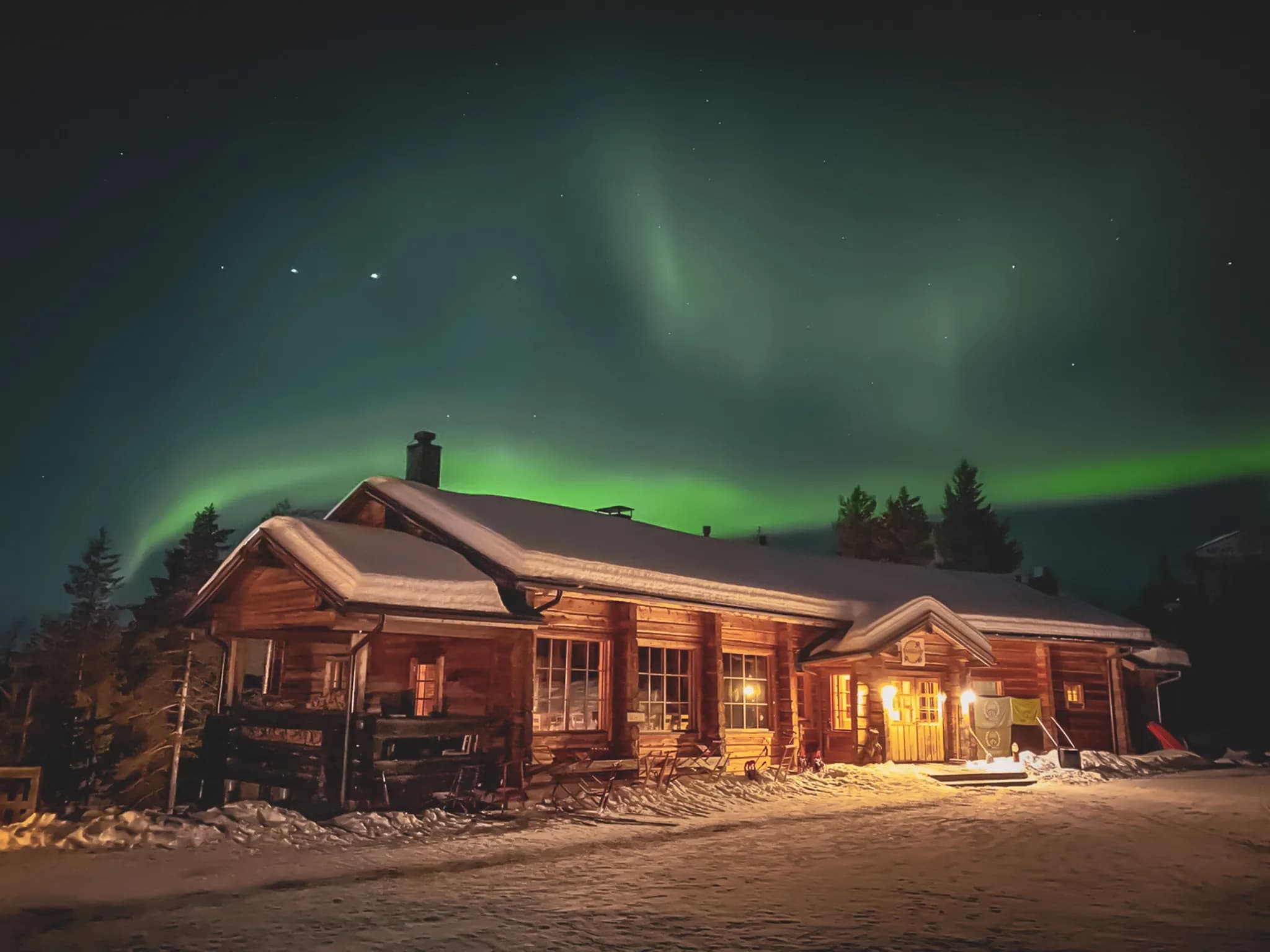 An illuminated wooden chalet nestled in a snow-covered landscape, under a night sky illuminated by green northern lights. The building has large windows that let in warm light,