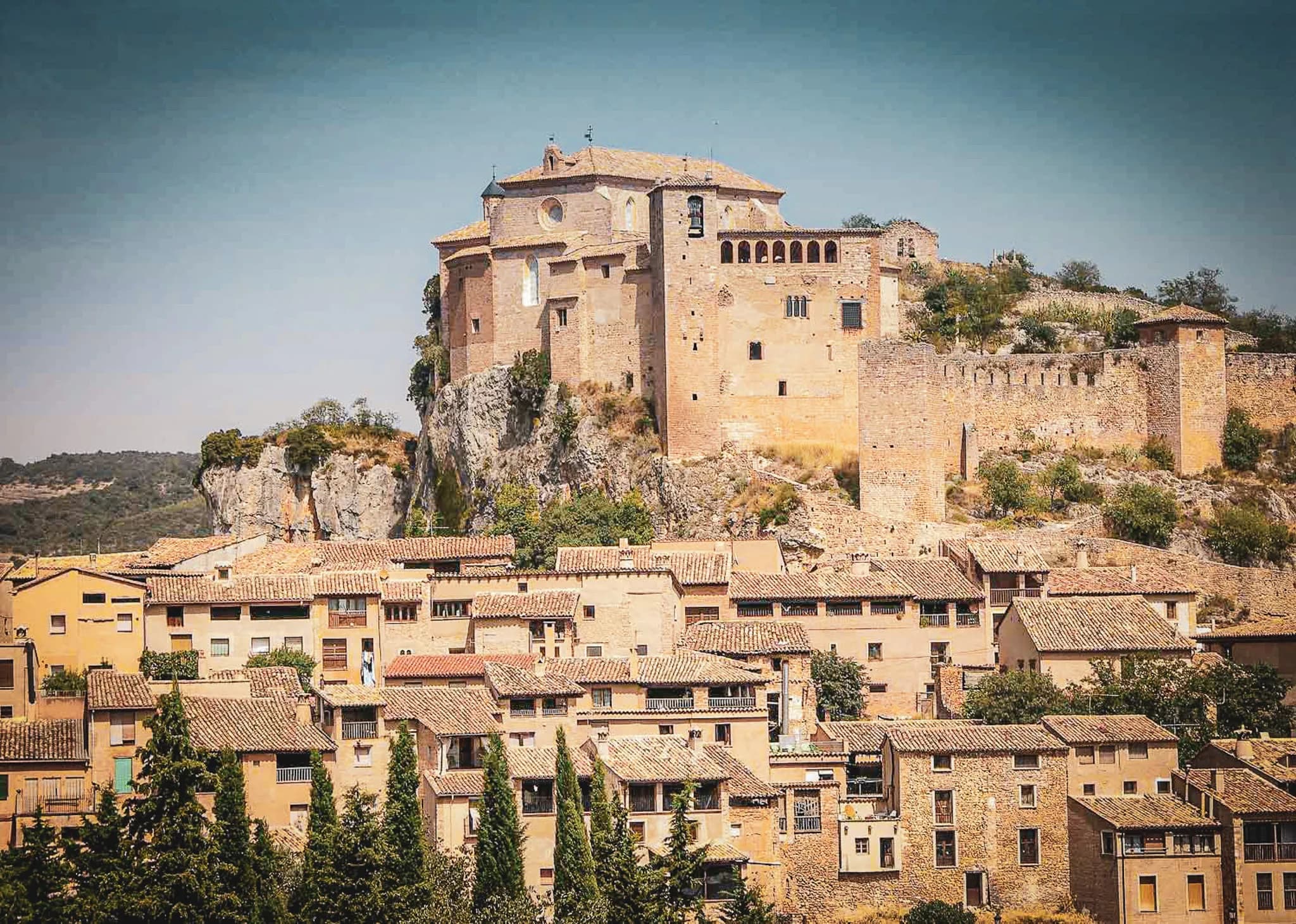 A medieval castle perched on a hill overlooks a picturesque village of houses with beige facades and tiled roofs. The castle, built of stone, features towers and windows of