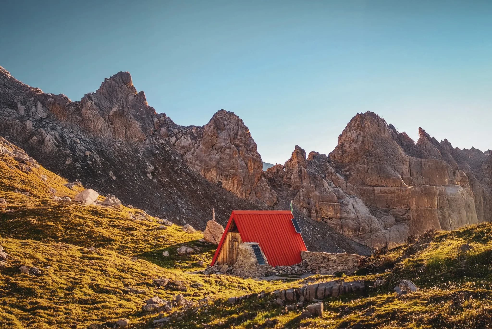 A chalet with stone walls and a red roof stands in the middle of an impressive mountain landscape. Steep, rock-riddled peaks surround the structure, while the surrounding terrain is recom