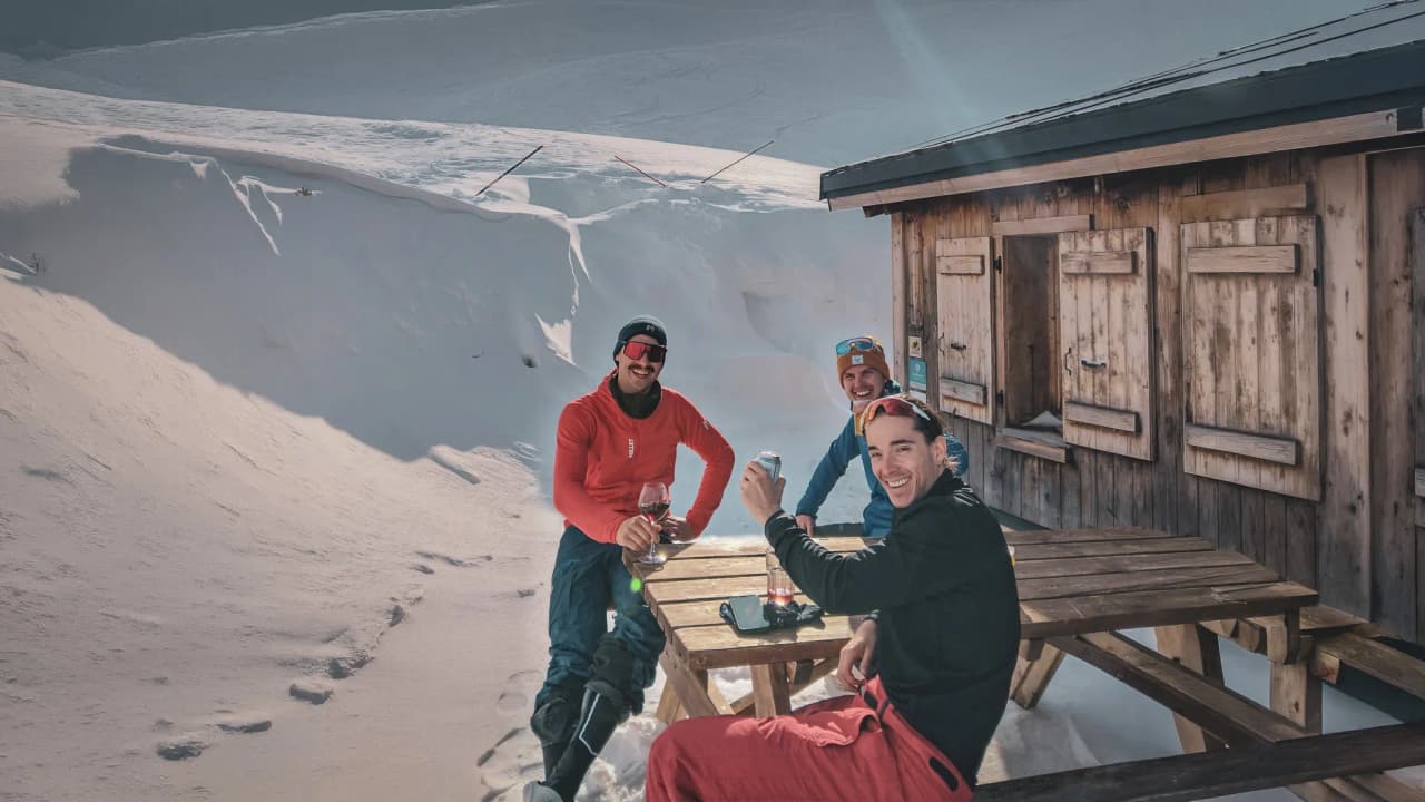 Three friends enjoy the sun and a drink in front of a mountain chalet, surrounded by snow.
