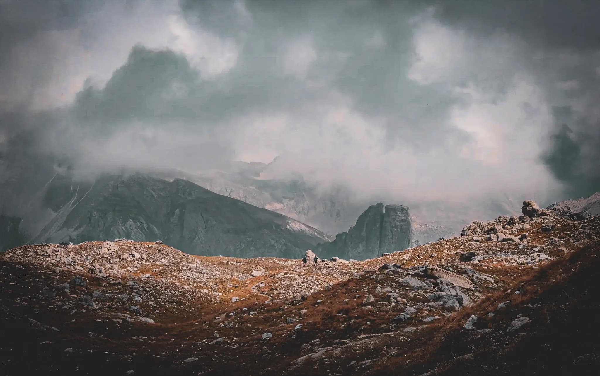A mountainous landscape shrouded in dark, heavy clouds, with craggy ridges emerging from the mist. The ground is strewn with rocks and sparse vegetation, creating a stark contrast.