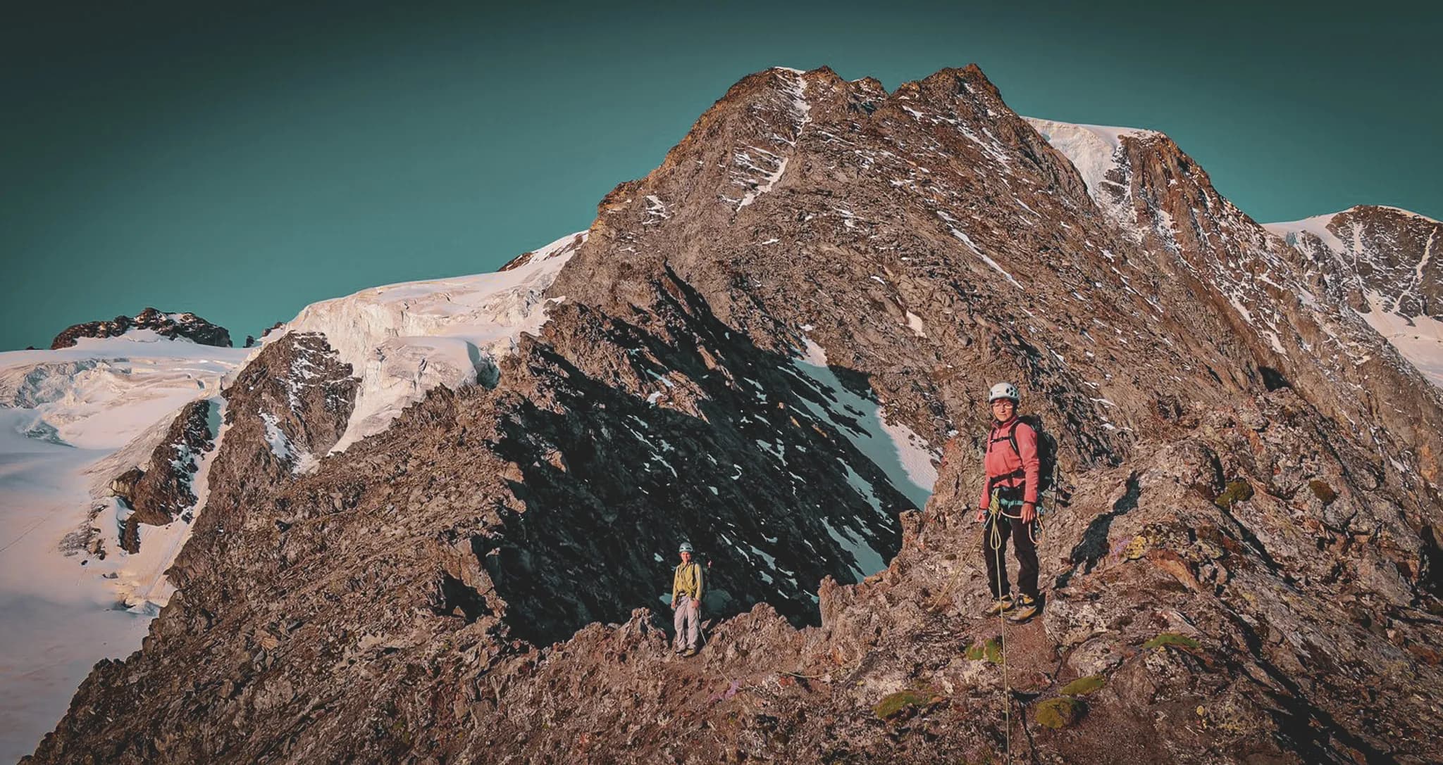 A majestic mountain landscape with two climbers standing on a rocky path. In the background, snow-capped peaks and glaciers sparkle under a clear blue sky. The mountains have walls