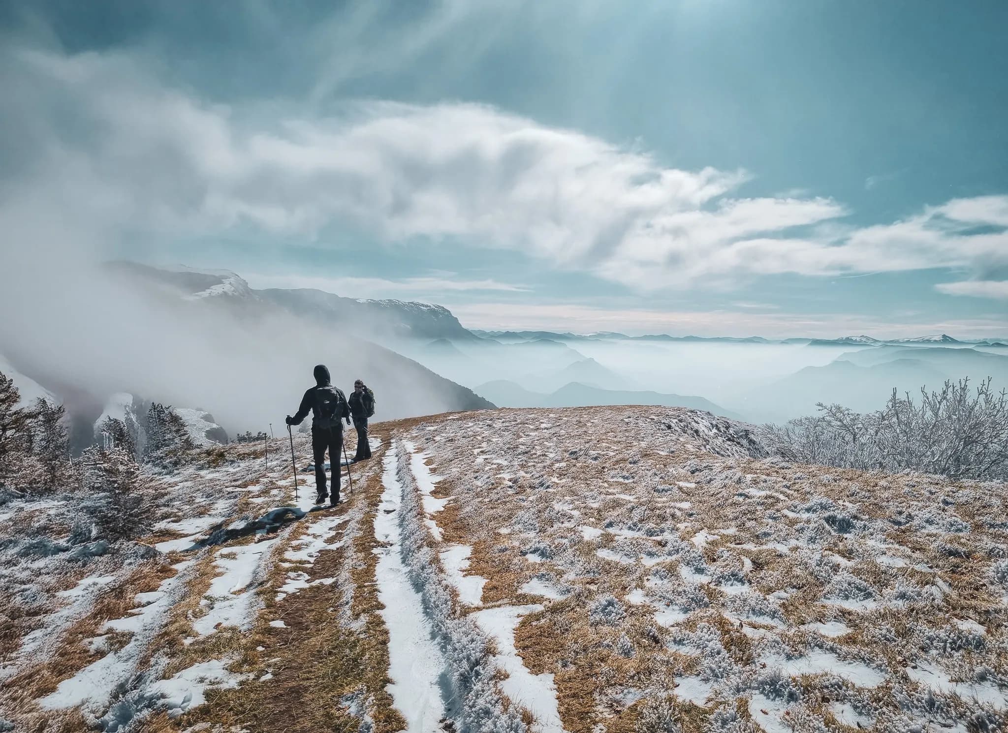 Two hikers walk along a snowy path, with majestic mountains in the background.