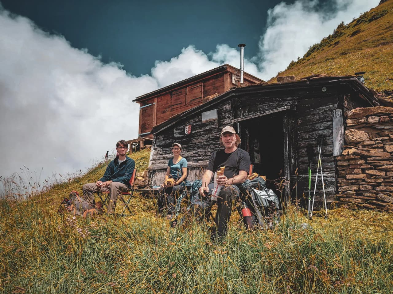 Three hikers pause in an Alpine landscape, surrounded by meadows and a rustic hut.