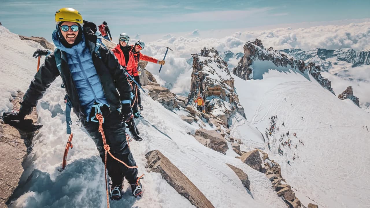 A group of smiling skiers on the snow-covered summit of Gran Paradiso, with a breathtaking panorama in the background.
