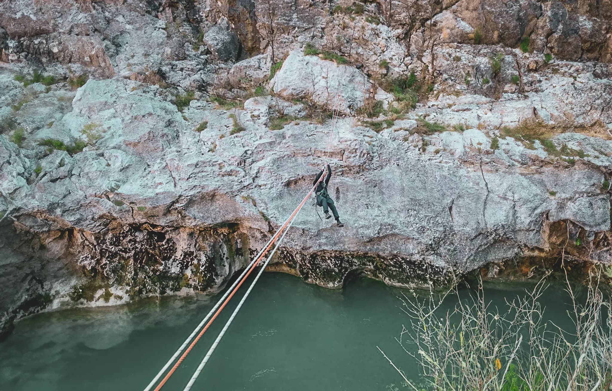 An adventurer suspended by a rope above a stream, abseiling down a rock face. The rocks, covered with moss and scattered grasses, create a