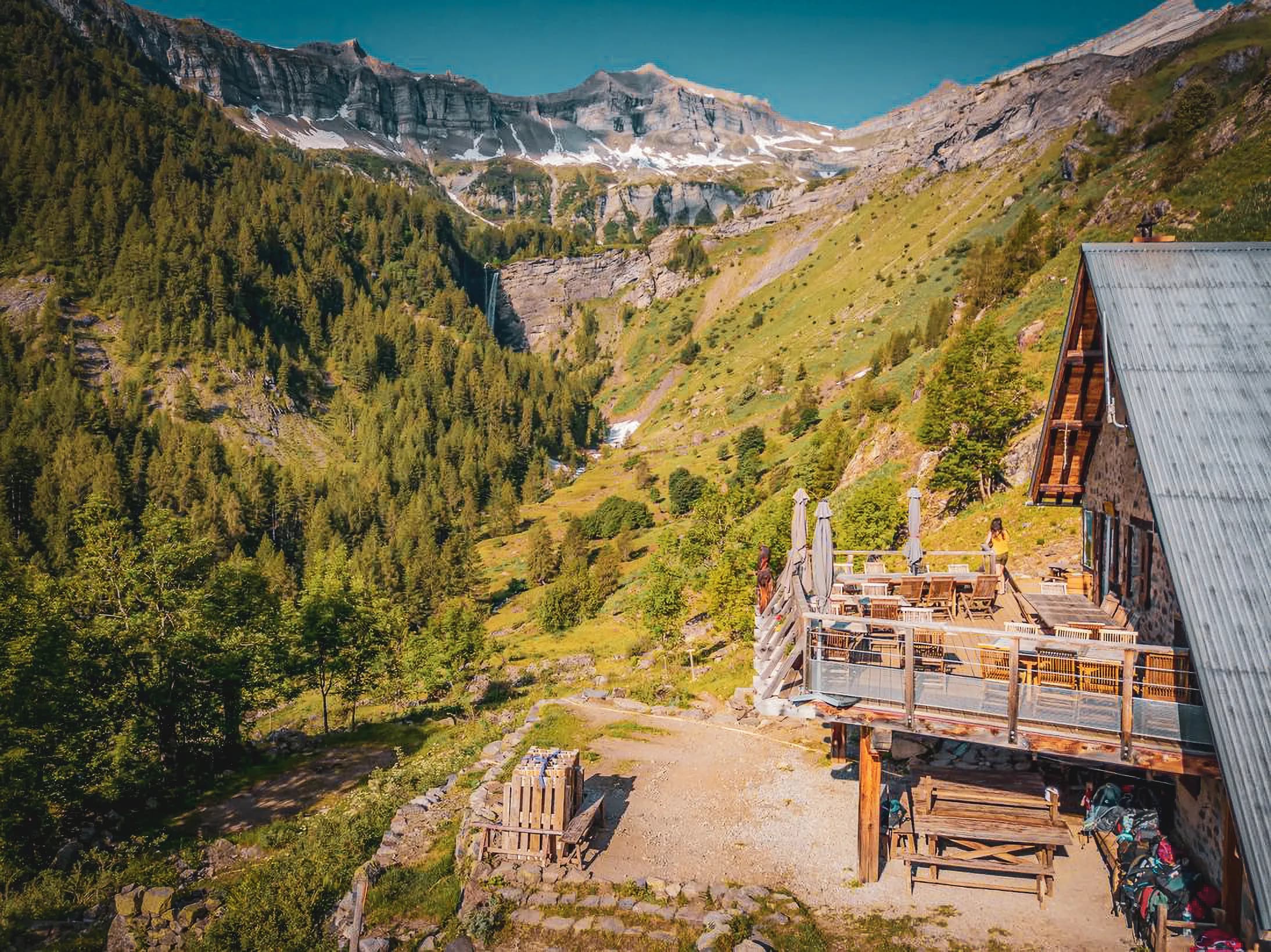 A wooden chalet at a high altitude overlooking a green valley. The chalet's terrace is furnished with tables and chairs, surrounded by parasols. In the background, you can see the mountains.