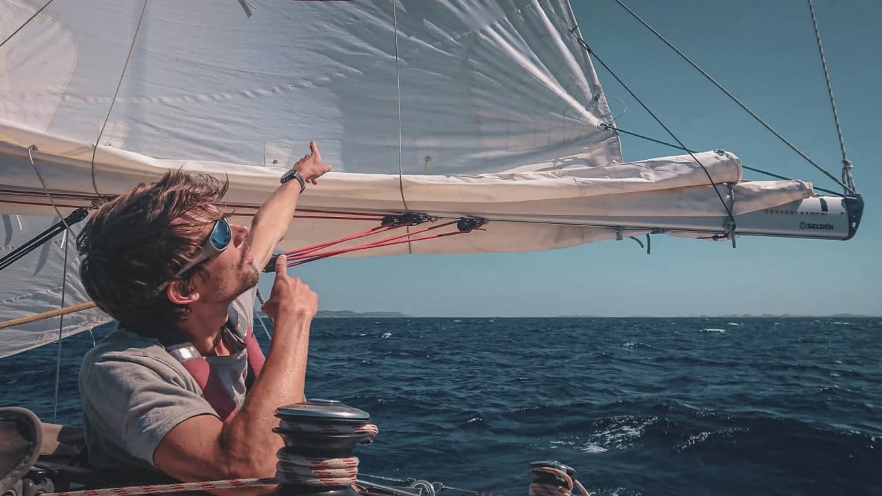 Un marin souriant, en pleine navigation, pointe vers les voiles sous un ciel ensoleillé.