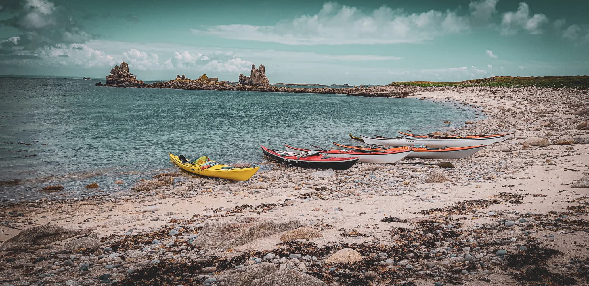 A coastal landscape with a pebble beach, where several brightly coloured kayaks, including a yellow one, are washed up on the shore. In the background, rock formations emerge from the sea, under a clear sky.