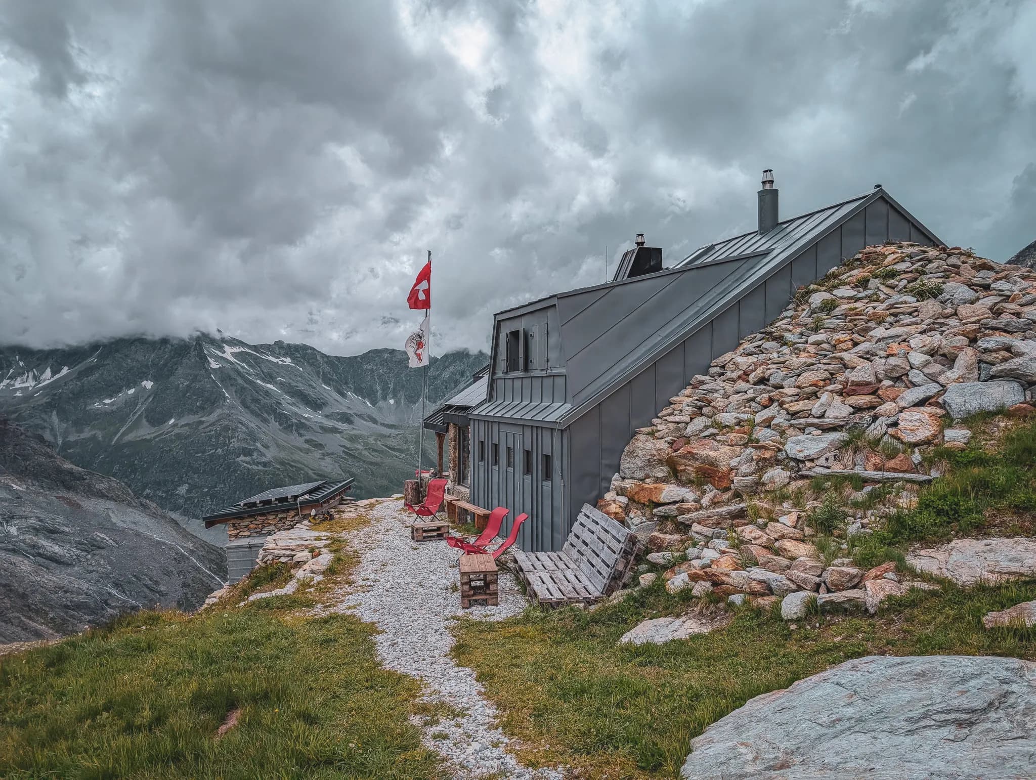 A modern building with a sloping roof is situated high up, surrounded by mountains. Two flags are visible: one is the Swiss flag, and the other bears a coat of arms.