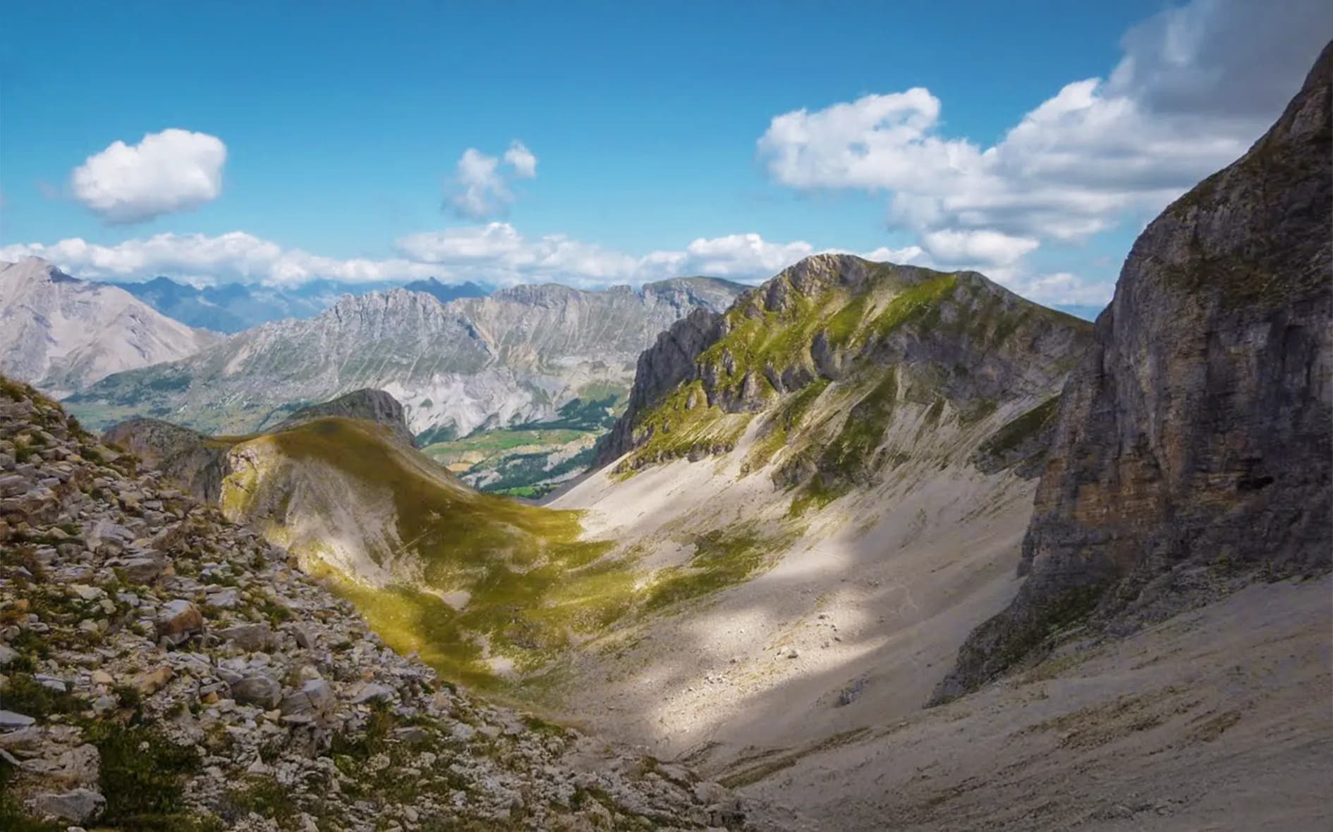 A vast mountain landscape with steep ridges and lush green valleys. The mountains are shades of grey with patches of green. The sky is light blue with white clouds,