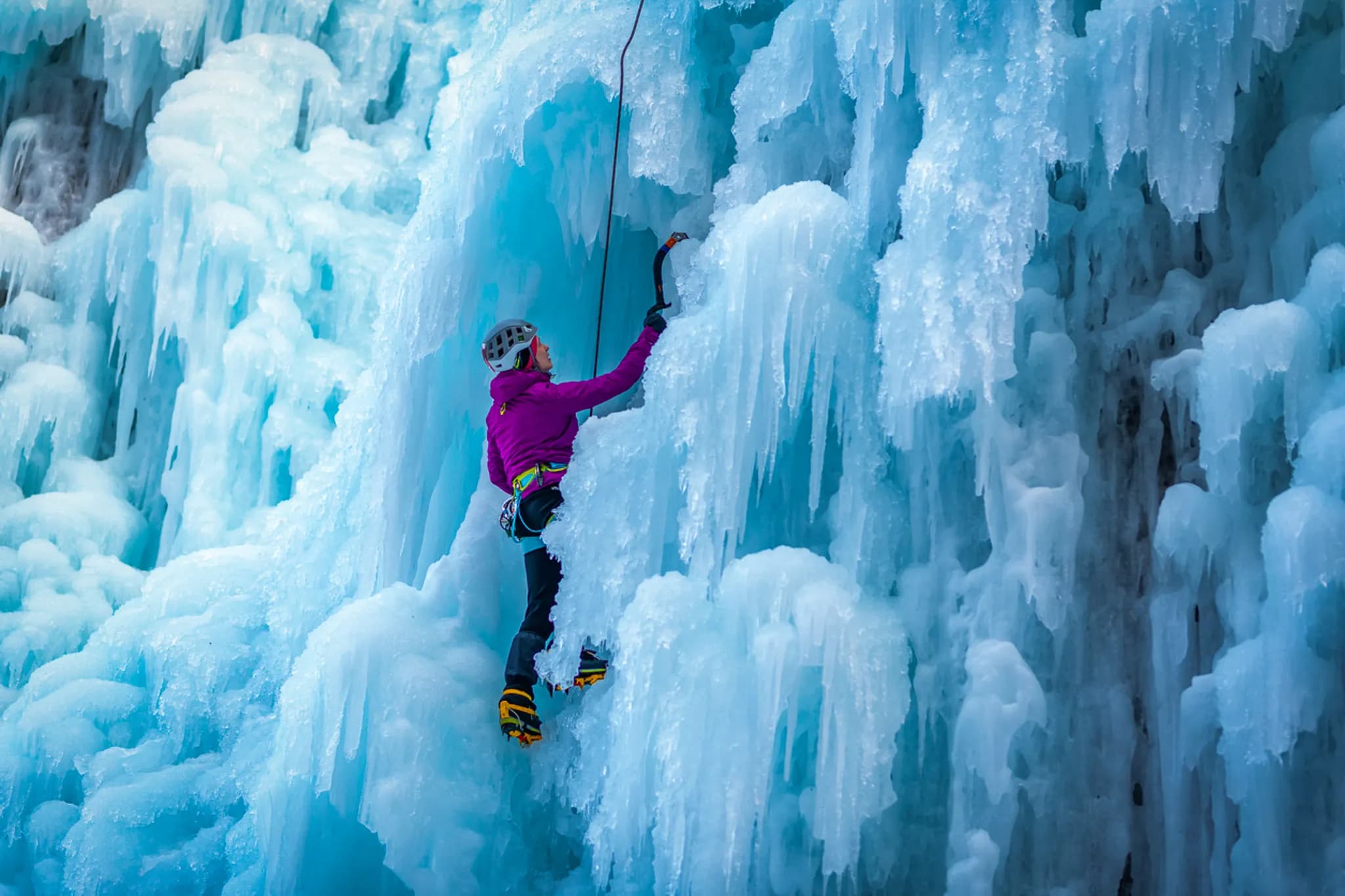 Un grimpeur escalade une imposante formation de glace, entouré de parois glacées d'un bleu clair et scintillant. Vêtu d'une veste rose et équipé d'un casque de sécurité, il utilise un piolet pour se