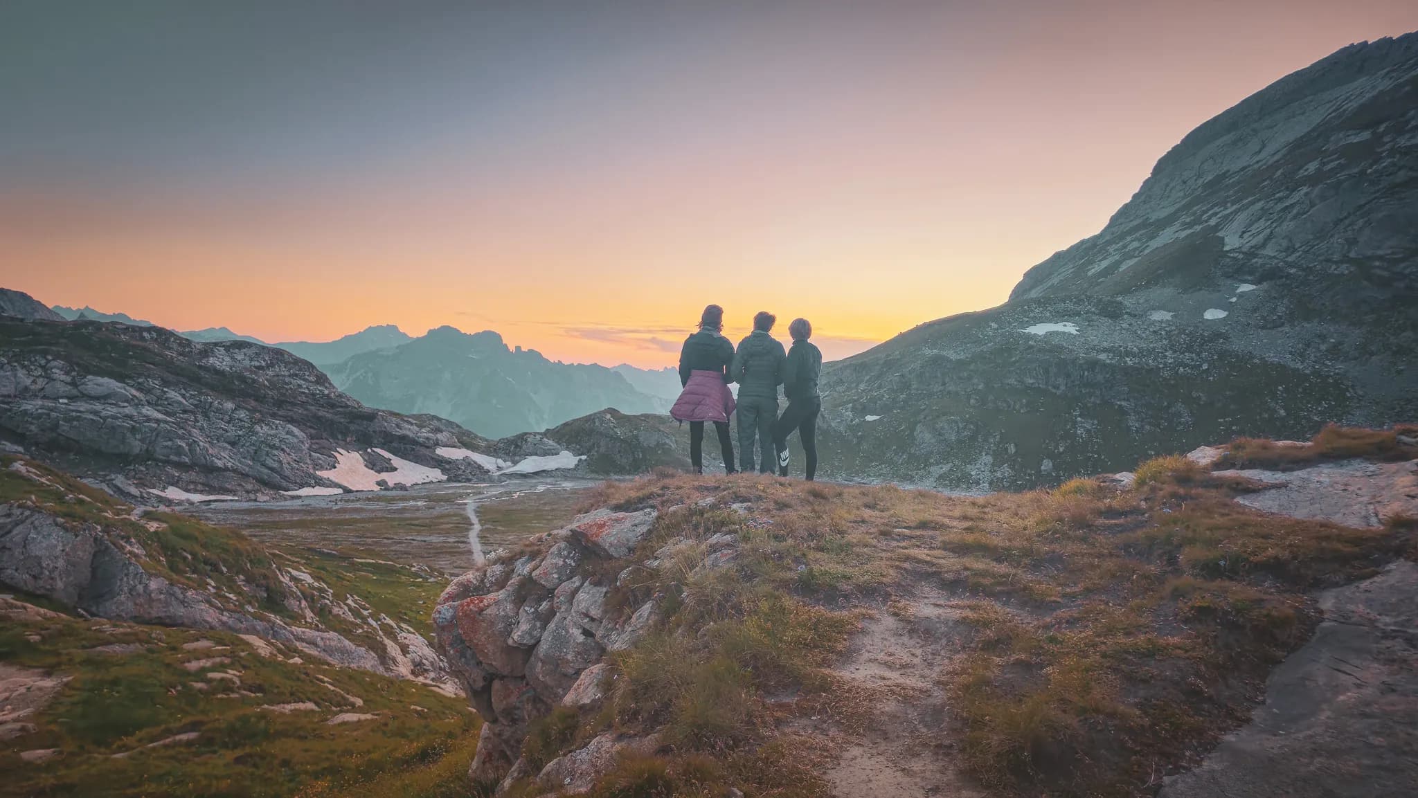 Un groupe de trois personnes se tient au bord d'une falaise, admirant un paysage montagneux au coucher du soleil. Les silhouettes des montagnes se dessinent en arrière-plan, baignées dans des teintes chaudes de rose et d'orange