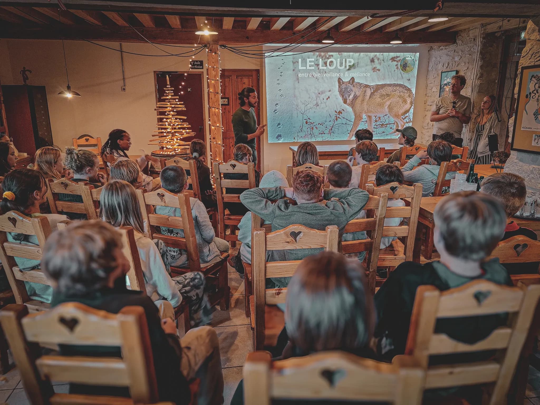 A presentation takes place in a room where a group of children sit on wooden chairs with carved hearts. At the front, a screen displays an image of a wolf, accompanied by the title "LE LO