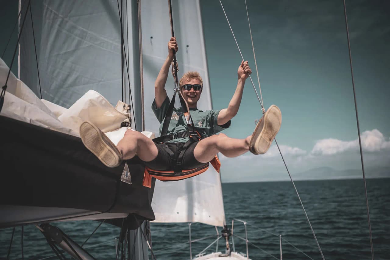 Un jeune homme souriant, suspendu à un voilier, sur des eaux méditerranéennes scintillantes.