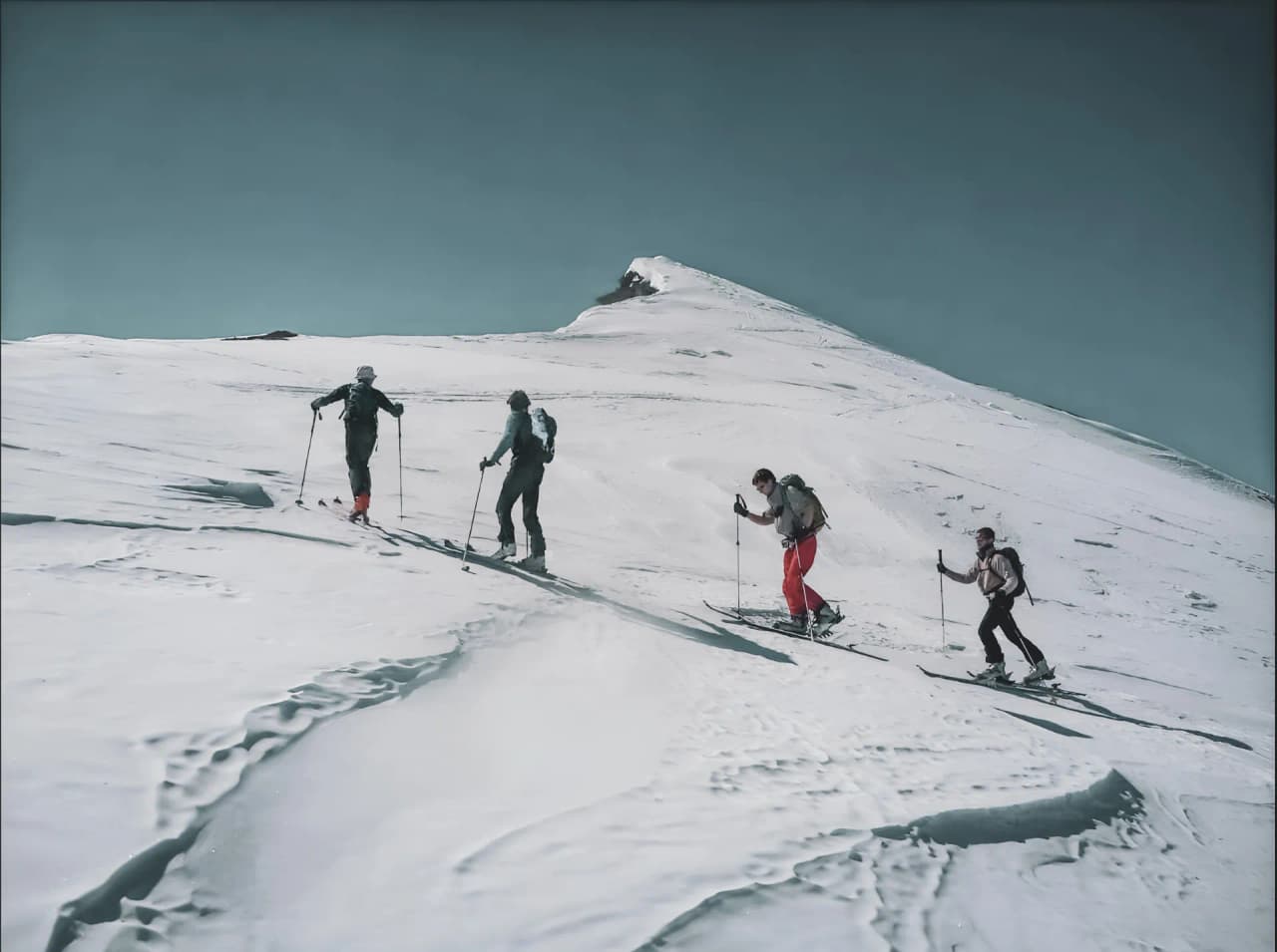 Quatre aventuriers en ski de randonnée gravissent des pentes enneigées, sous un ciel bleu éclatant.