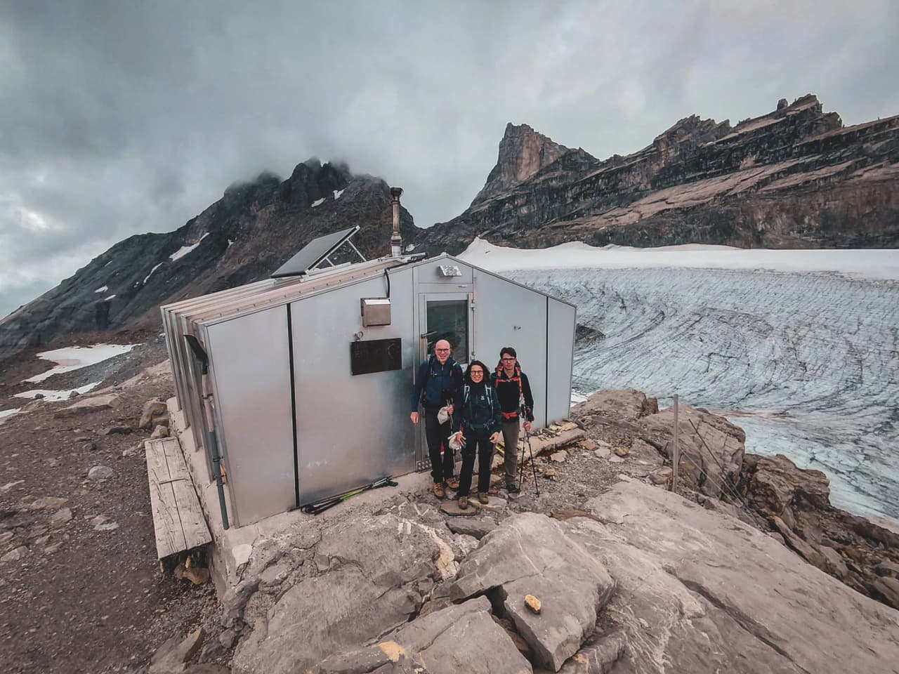Three hikers pose in front of an alpine Mountain hut, with majestic mountains in the background.