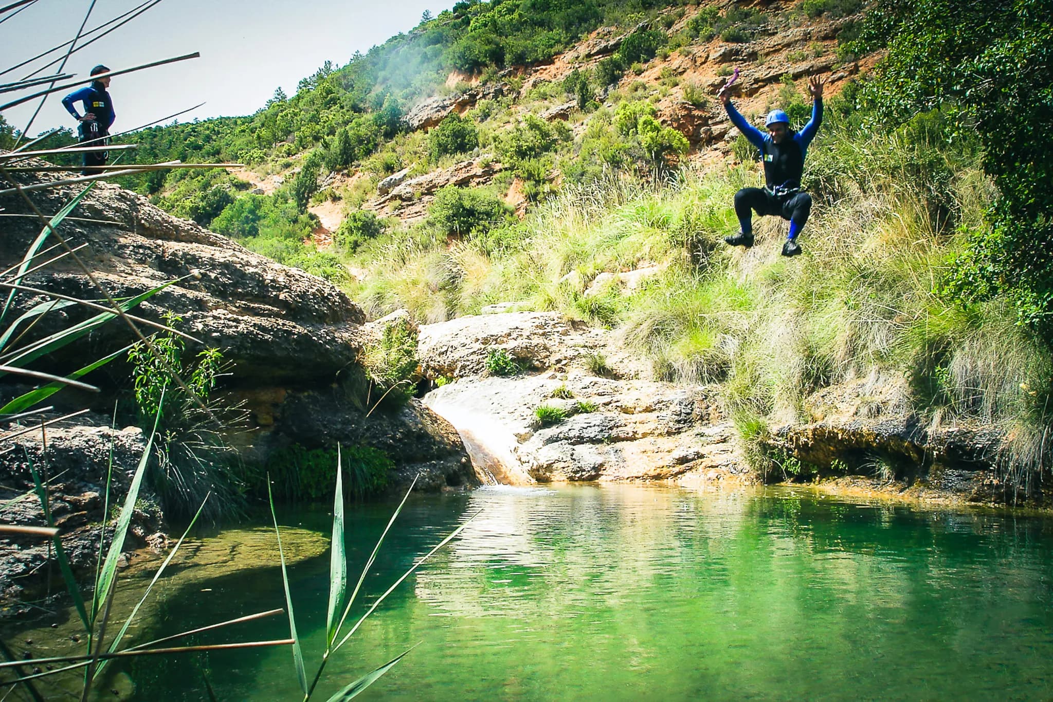 An adventurer in a diving suit makes a jump from a rock overlooking a natural pool, surrounded by lush vegetation. In the background, another participant stands on a rock, looking out over the pool.