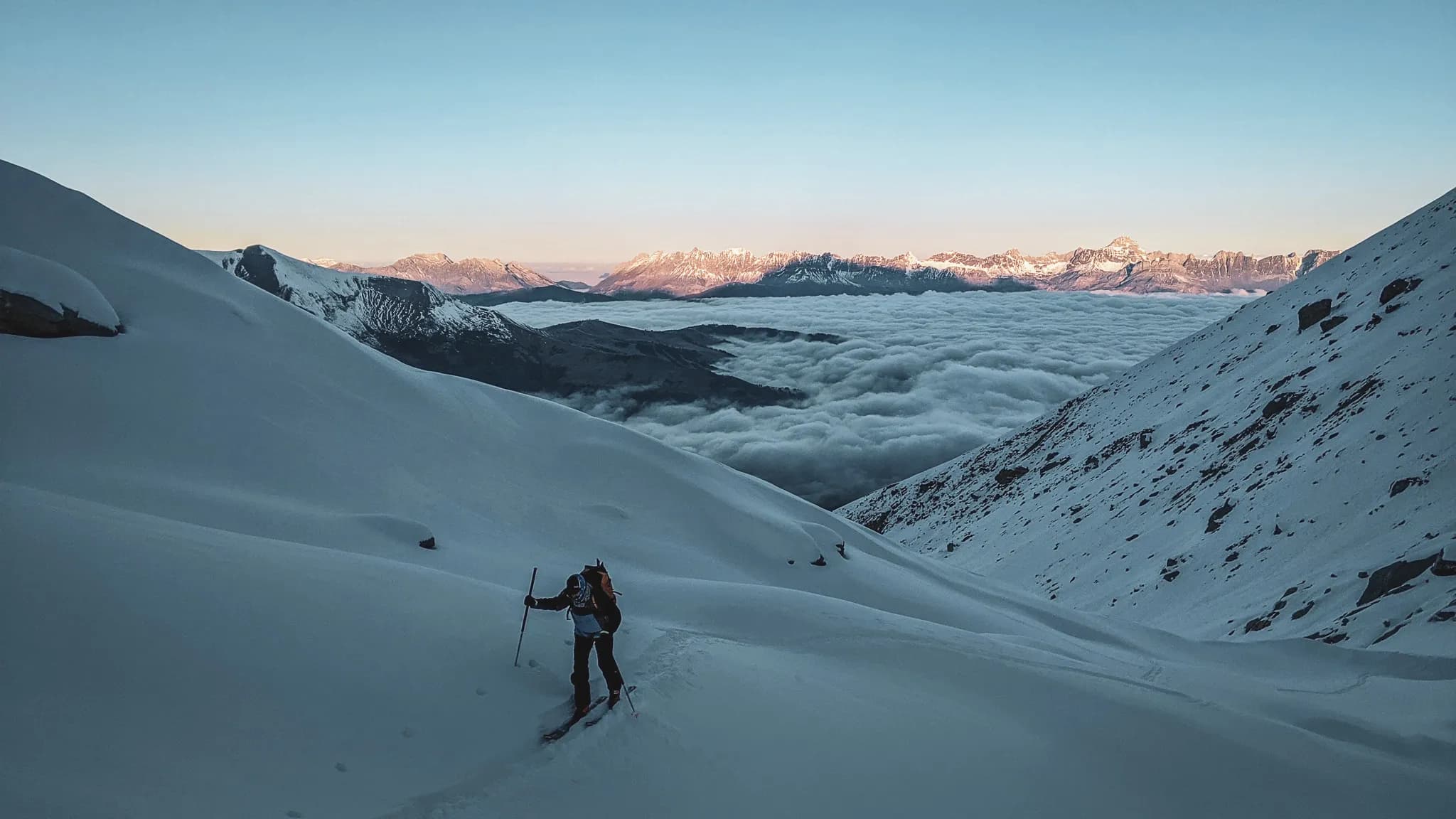 Un skieur évolue sur des pentes enneigées, entouré de vastes paysages montagnards. En arrière-plan, une mer de nuages s'étend sur la vallée, tandis qu'à l'horizon, des sommets montagne