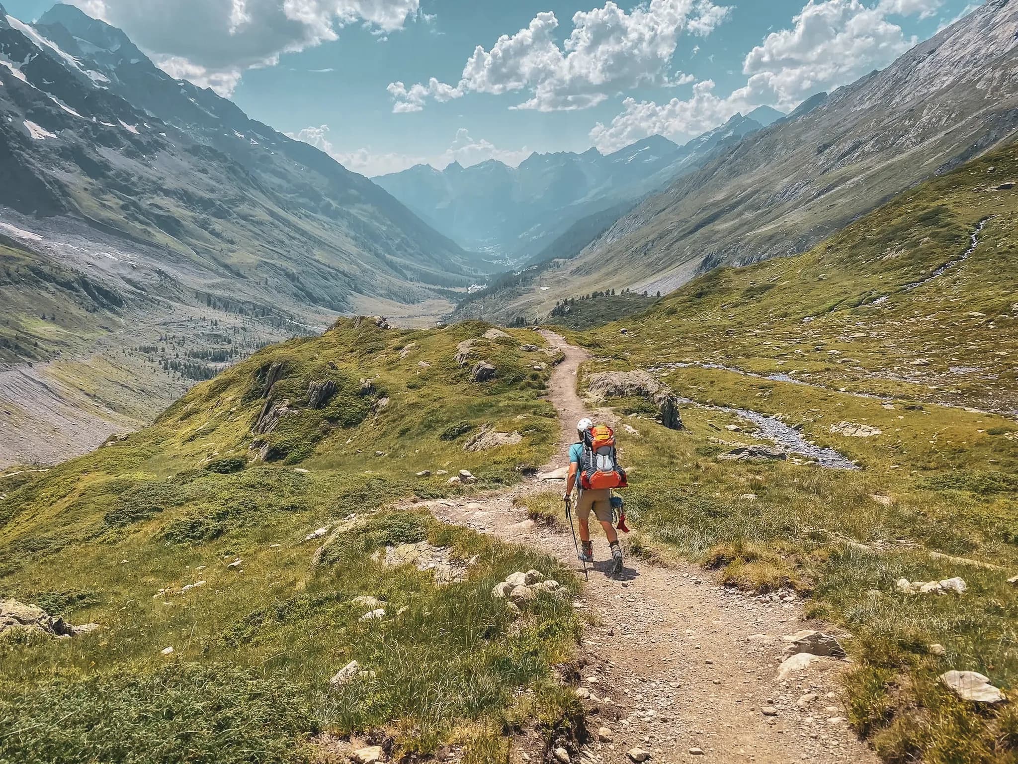 A hiking trail in the Falferlap valley winding through an Alpine landscape. Majestic mountains rise up in the background, with snow-capped peaks and lush green valleys.