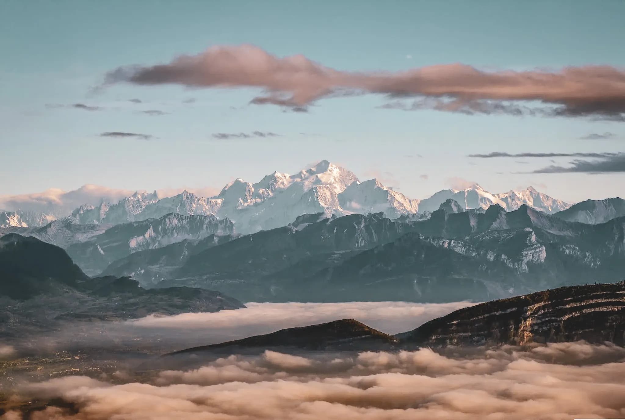 A majestic mountain landscape, with snow-capped peaks standing out against a clear blue sky. In the foreground, verdant, woody trees add a touch of wilderness, while a light mist