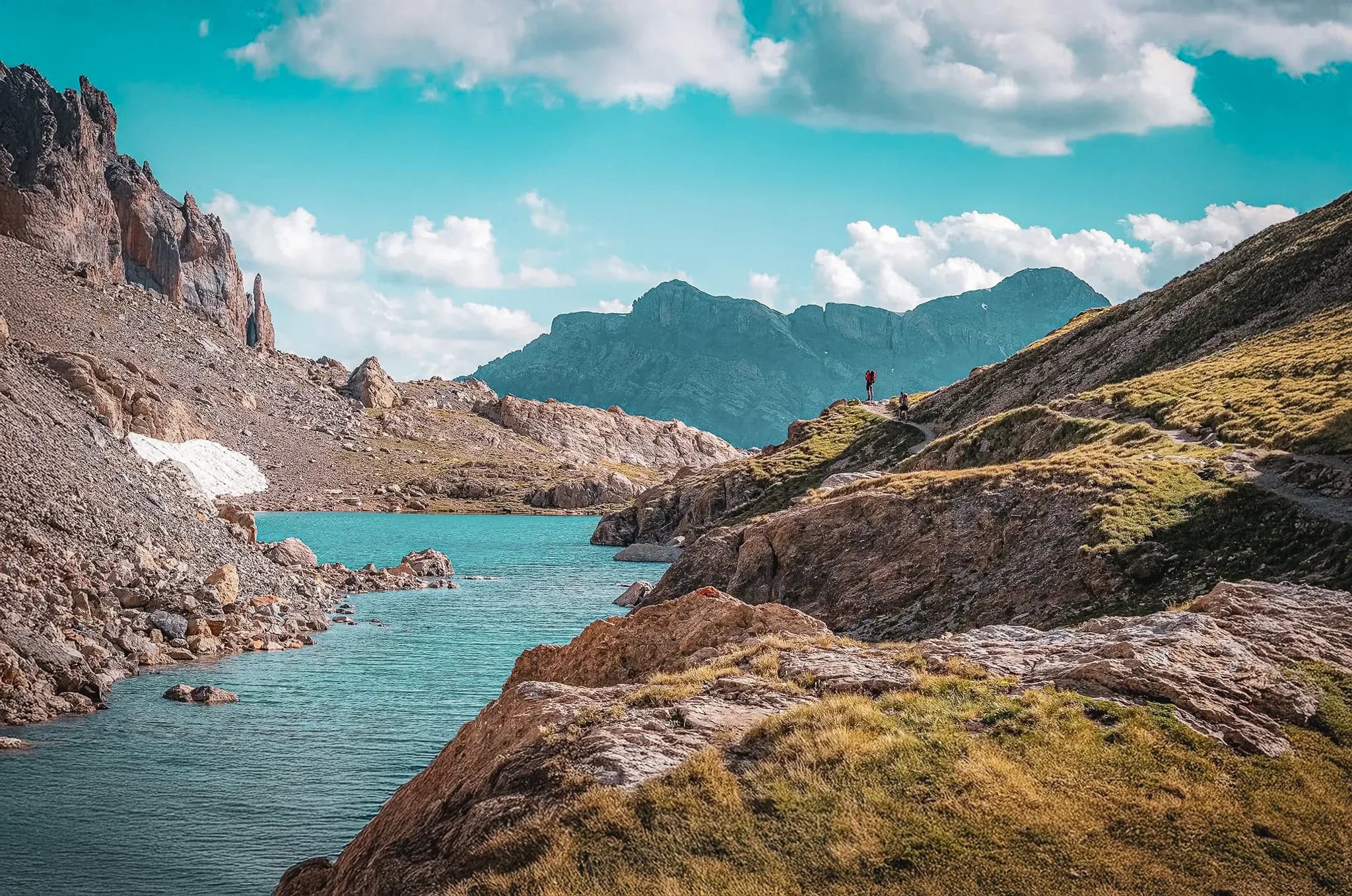 A mountain landscape with a turquoise lake surrounded by rock formations. Green slopes rise to steep peaks, under a clear sky dotted with white clouds. On the horizon, silhouettes