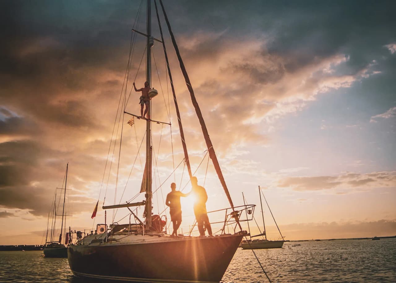 A sailboat illuminated by the sunset, sailors manoeuvring enthusiastically at sea.