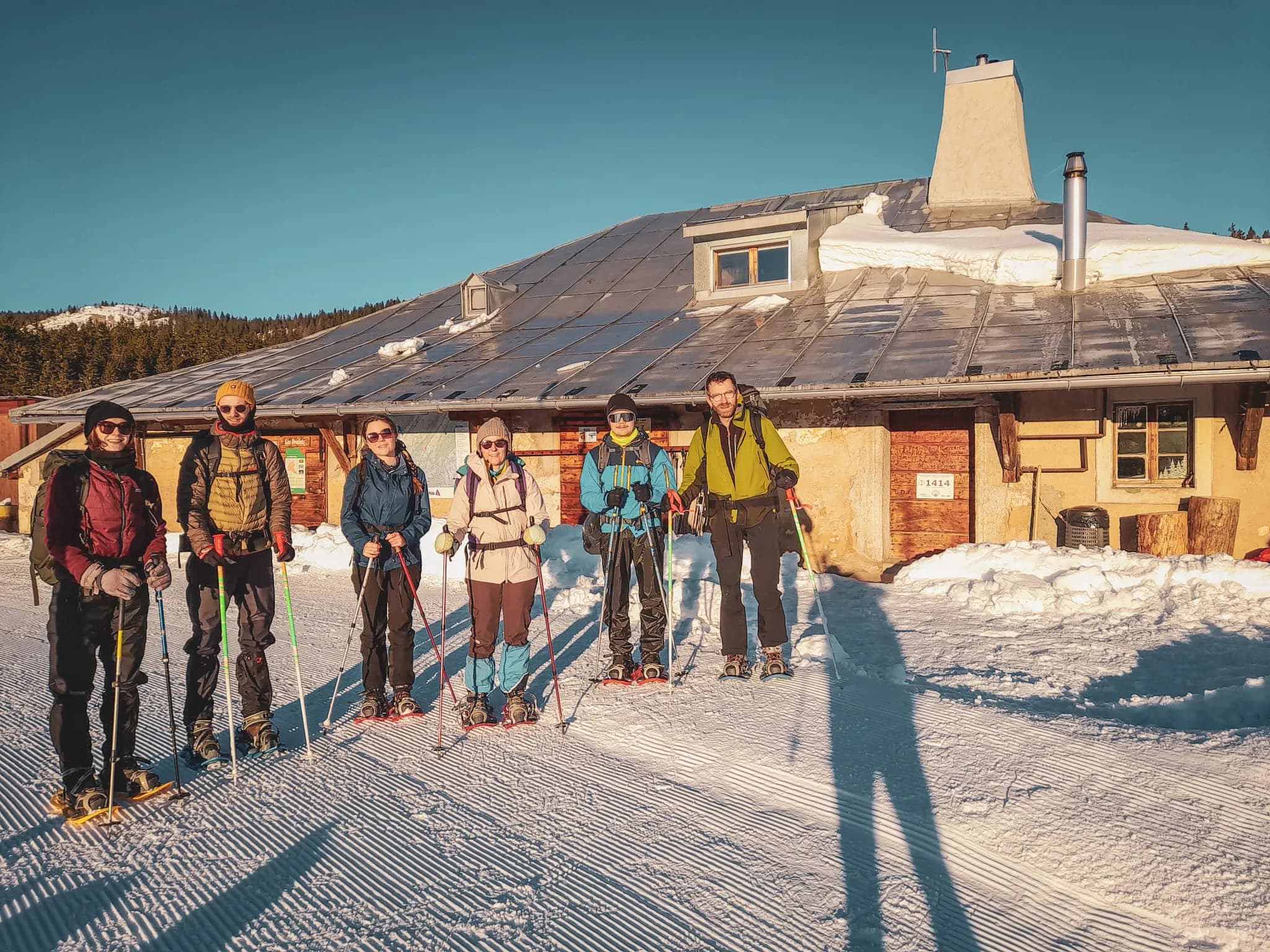 Een lachende groep op sneeuwschoenen voor een chalet in de zon, klaar voor hun winteravontuur.