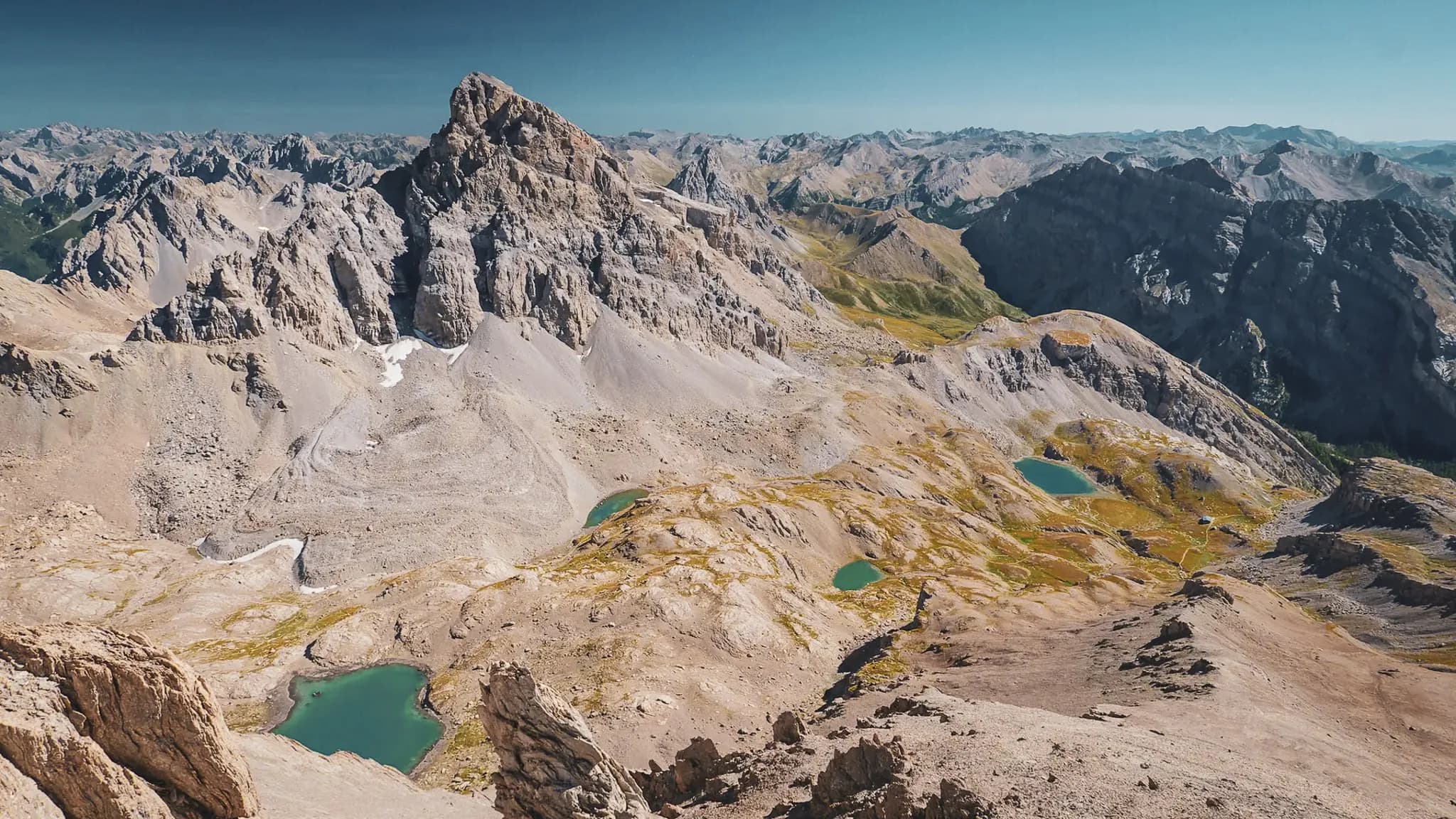 A majestic mountain panorama of craggy, rocky peaks, lush green hills and turquoise lakes scattered across the valleys. The sky is a deep, cloudless blue, bringing