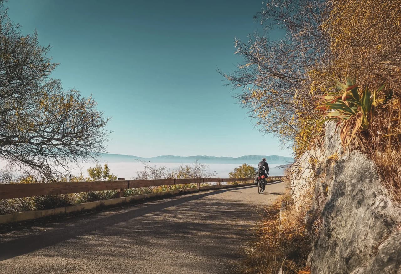 A cyclist pedals along a winding road in Italy, overlooking a misty valley. Trees with golden leaves line the path, while a wooden fence marks the boundary of the road. In the background