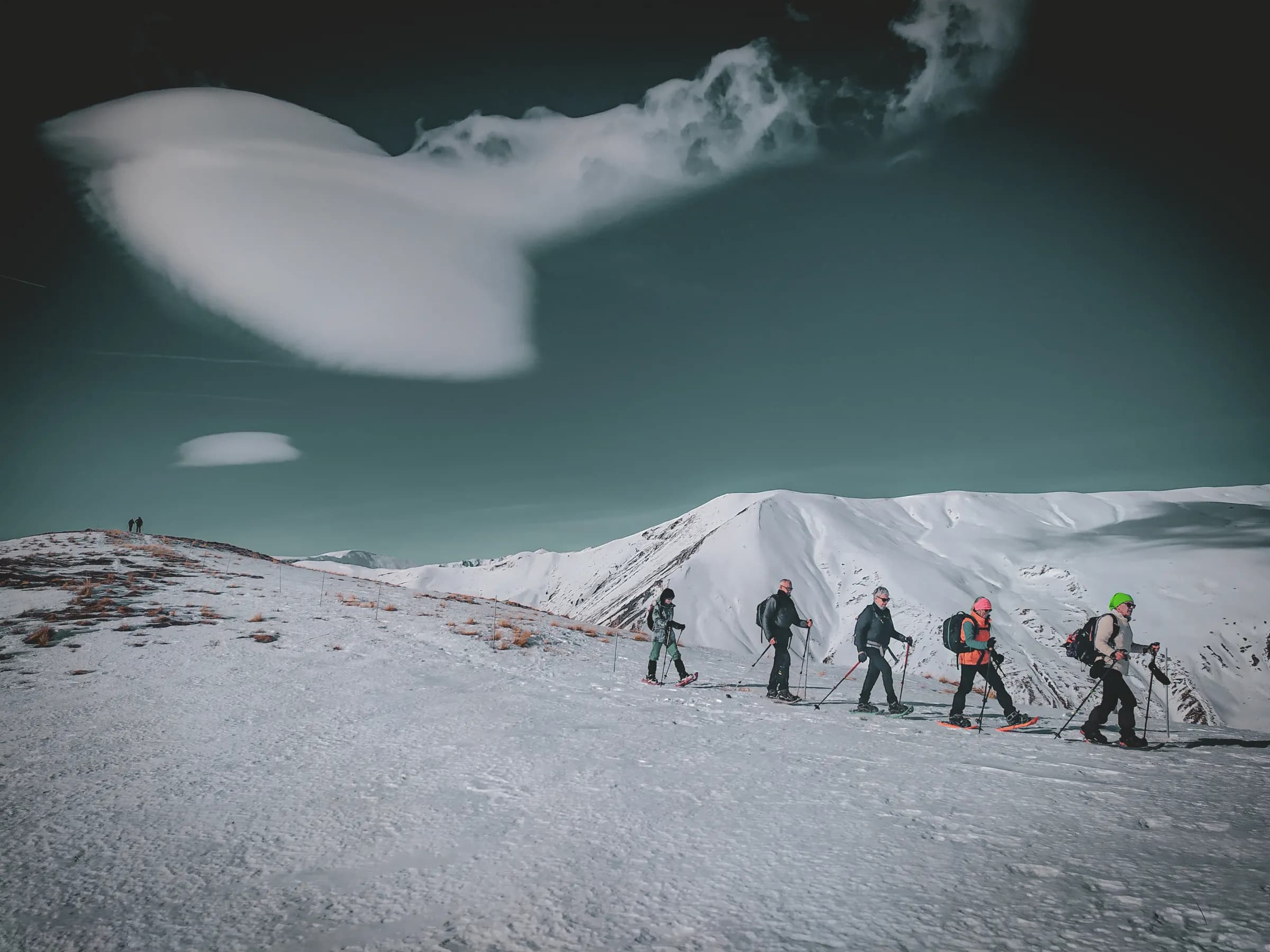 A group of hikers walk across snow-covered terrain, surrounded by majestic mountains under clear skies. The hikers are equipped with ski gear, some wearing helmets and colourful clothing.