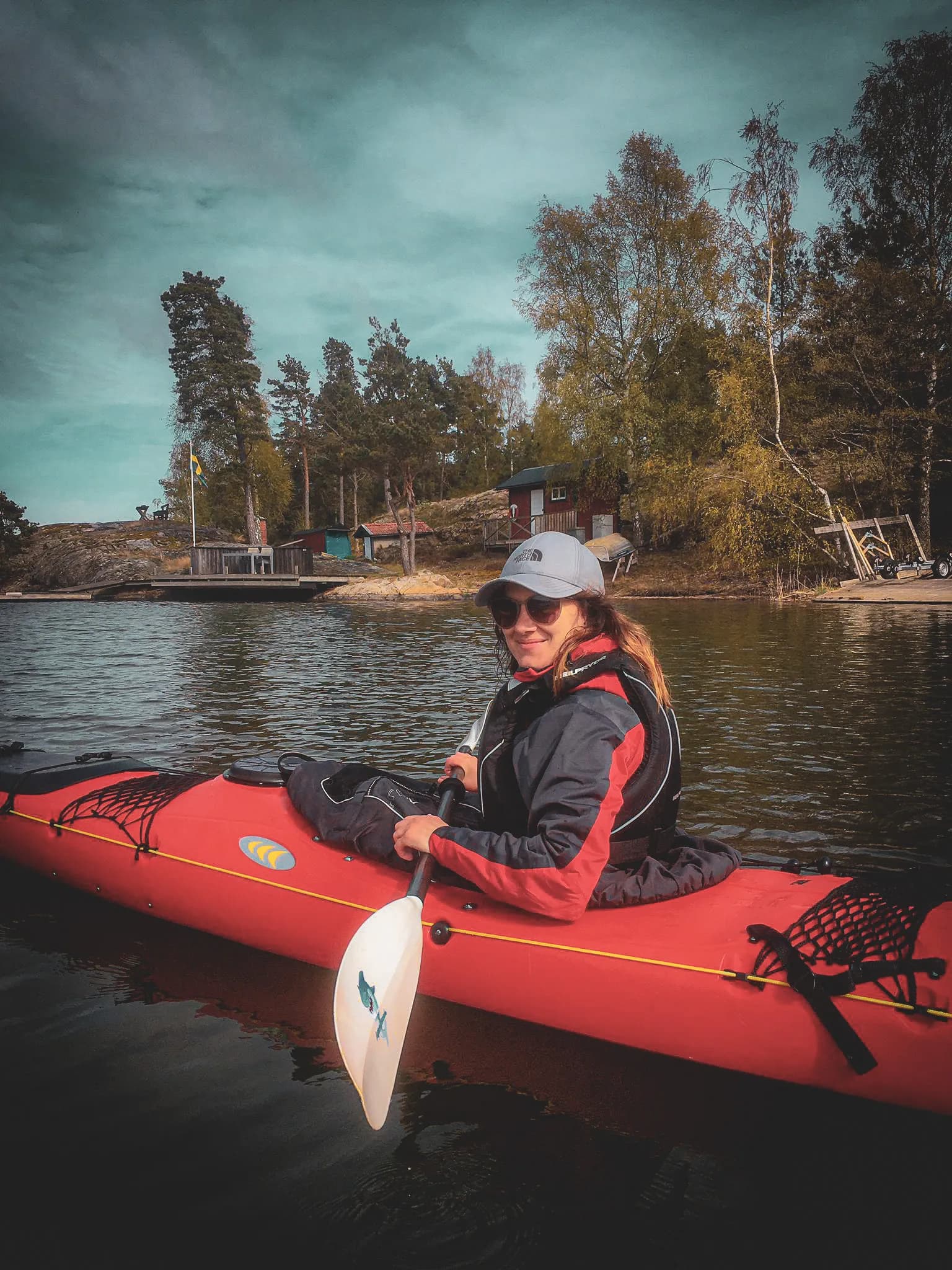 A young woman in a red kayak, smiling and wearing appropriate sportswear. She is wearing a grey cap and sunglasses. In the background, green trees and huts can be seen.